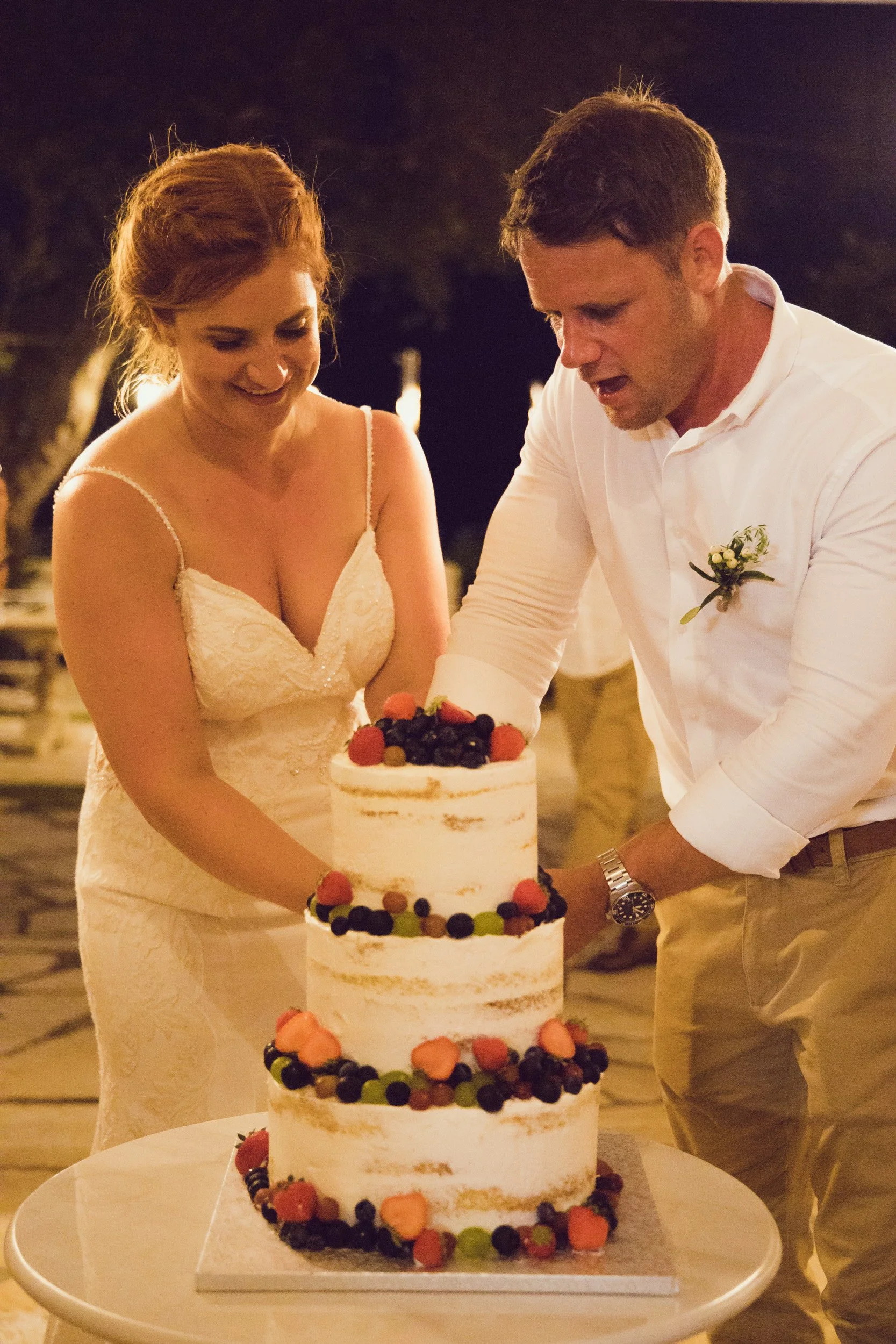 A bride and groom cutting a three-tier naked wedding cake decorated with berries and fruit during their outdoor reception at night.