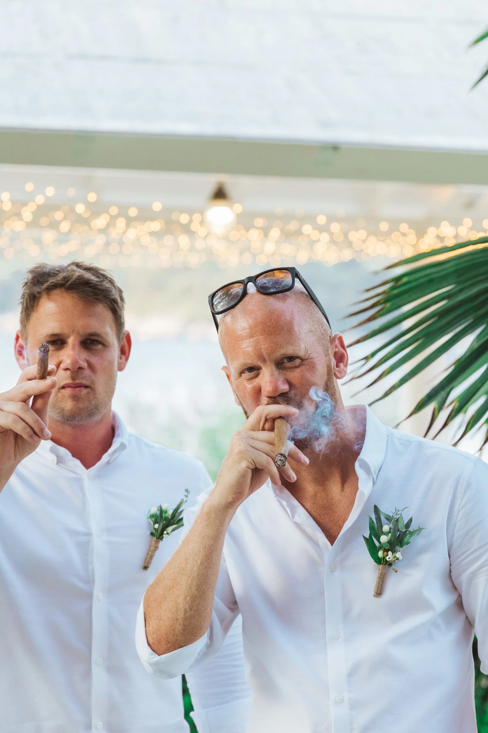 Two men in white shirts with boutonnières, smoking cigars, at an indoor event with string lights and tropical plants.