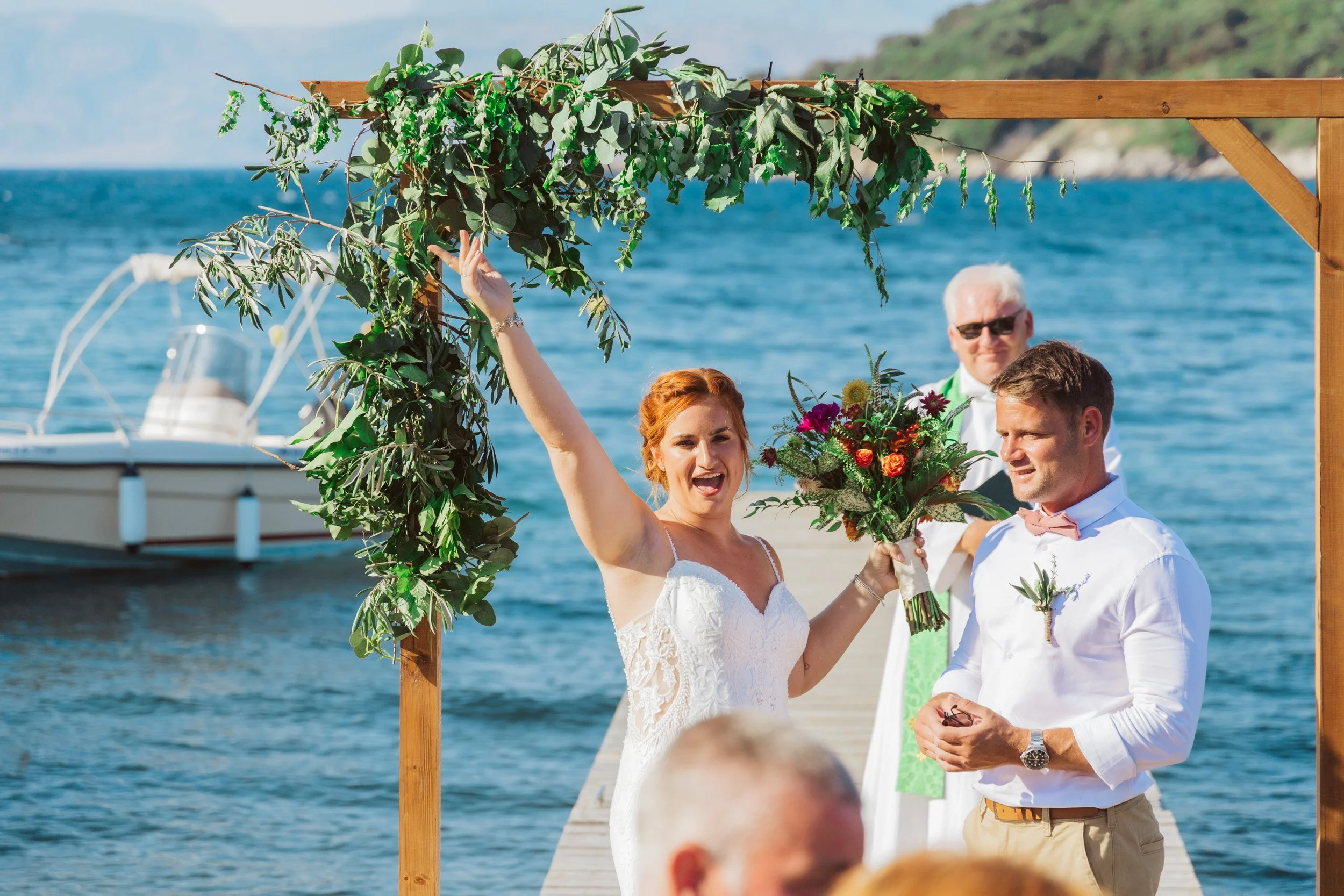 A couple getting married at a seaside wedding ceremony, with a woman in a wedding dress holding a bouquet and raising her hand, and a man in a white shirt with a pink bow tie. A priest or officiant stands behind them, and the ceremony is taking place