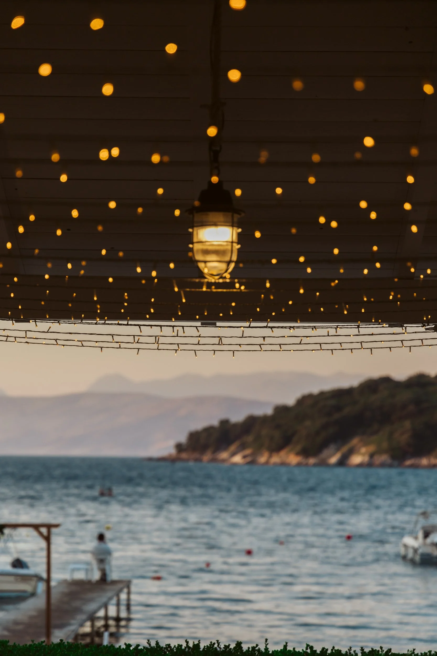 Outdoor scene with a hanging lantern and string lights, overlooking a body of water with boats and distant hills in the background during sunset.