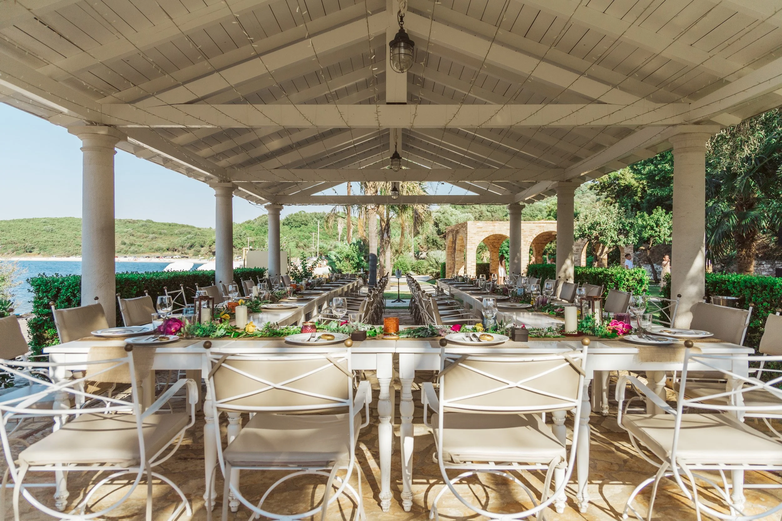An outdoor dining setup with a long white table under a covered pavilion, decorated with flowers, candles, and glassware, overlooking lush greenery and water in the background.
