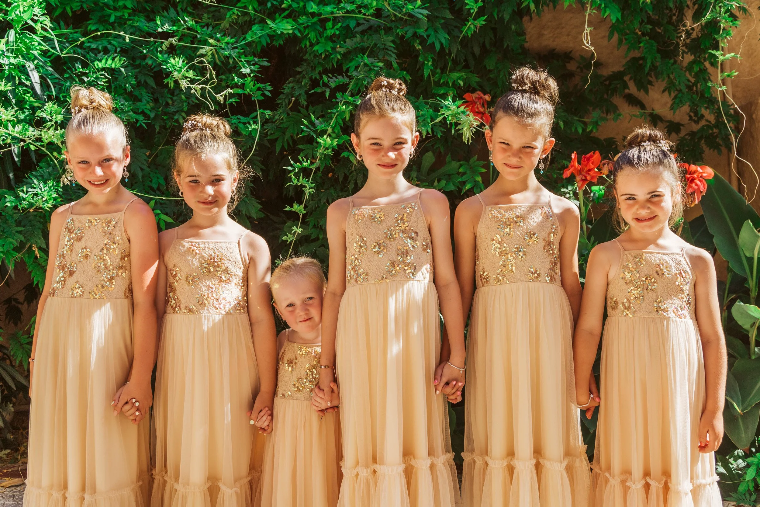 Seven young girls dressed in matching beige dresses with sequined tops, standing outdoors in front of green foliage and red flowers, holding hands and smiling.