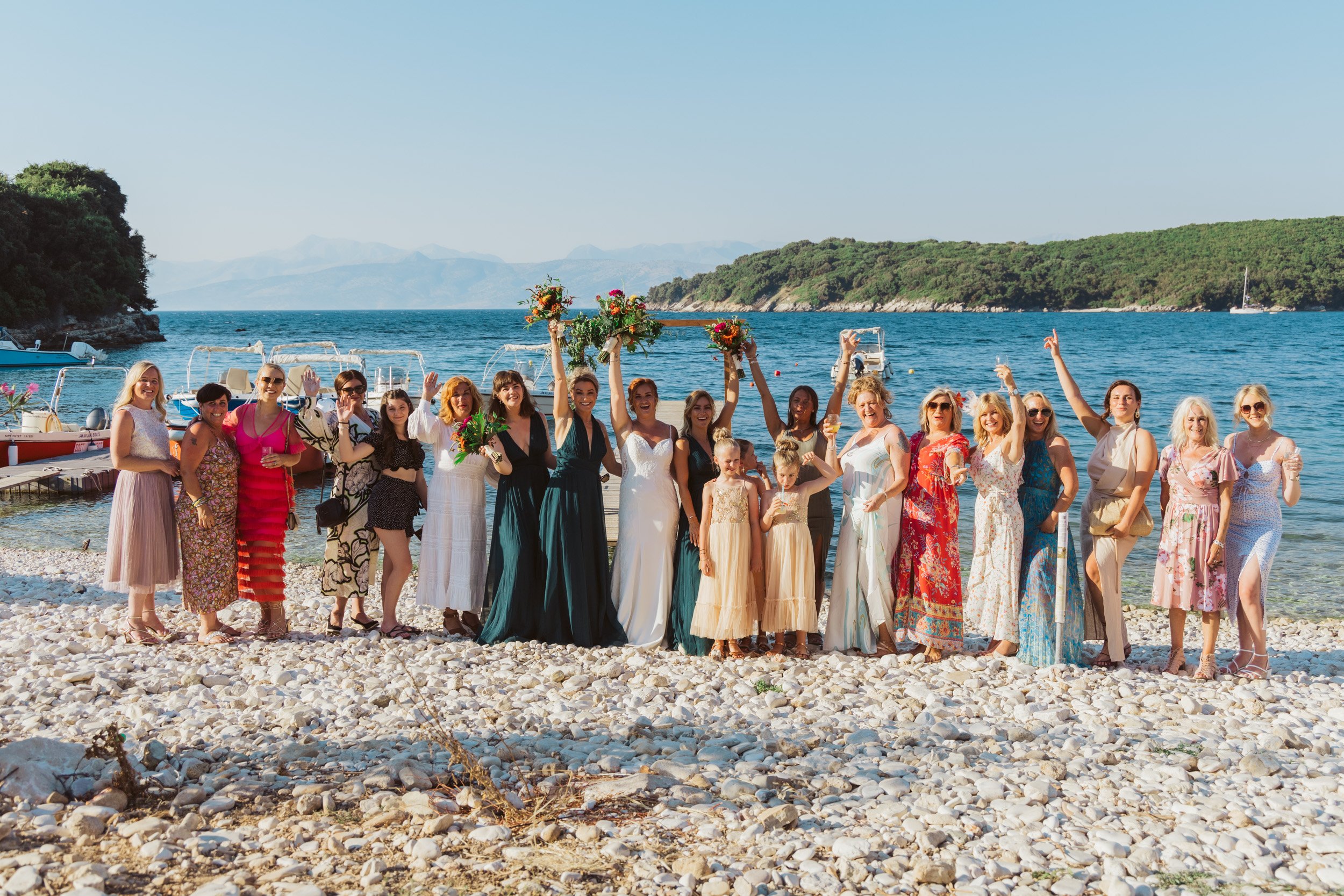 A group of women and children celebrating on a rocky beach near the water, some holding bouquets of flowers, with boats and distant mountains in the background.