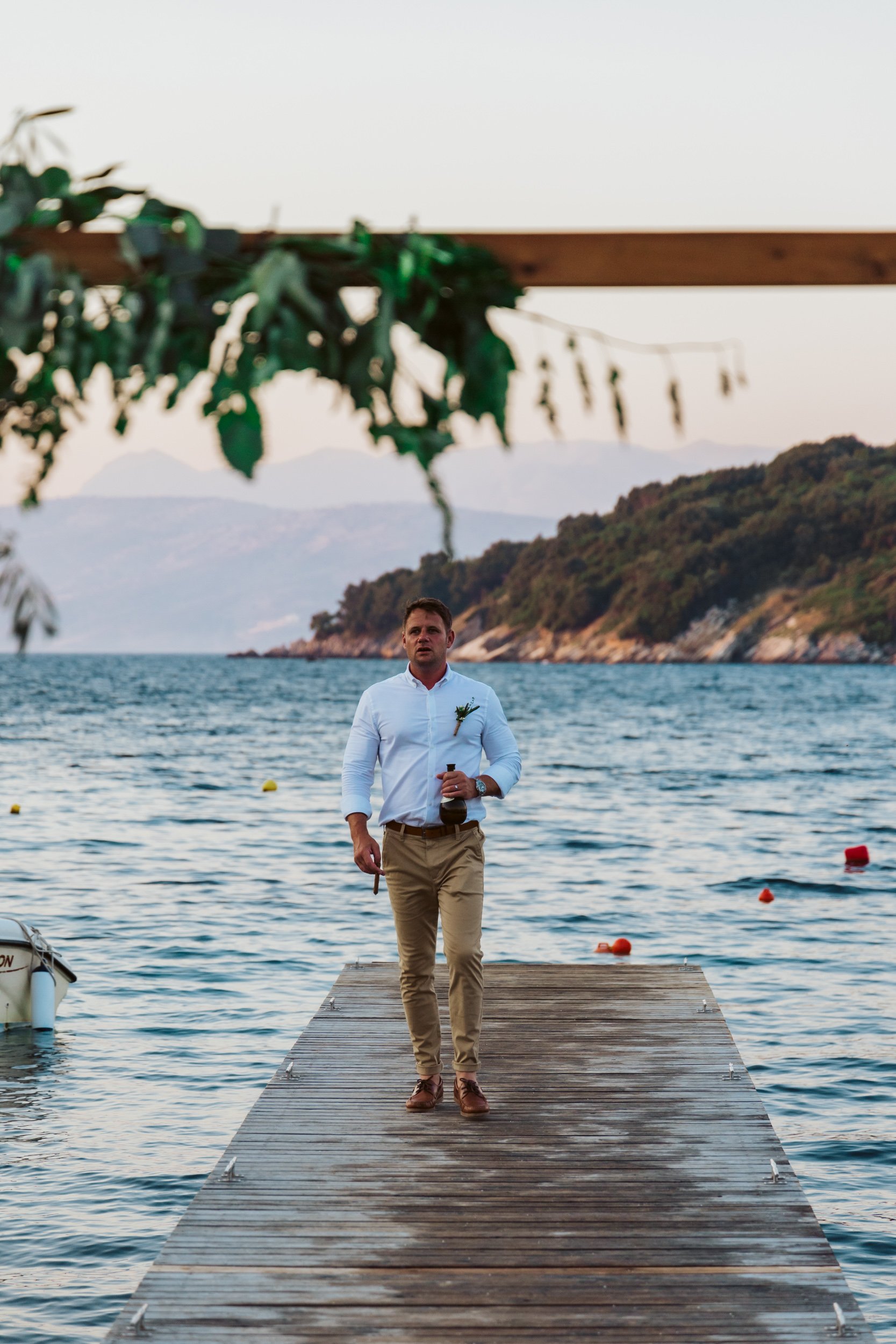 A man standing on a wooden dock by a lake during sunset, holding a drink, with boats anchored nearby and hills in the background.