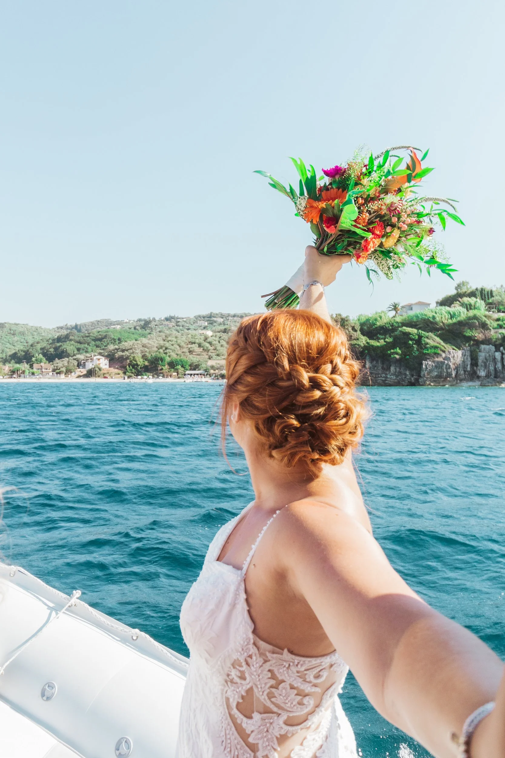 A woman with red hair styled in an updo, wearing a white lace dress, takes a selfie on a boat, holding a colorful bouquet of flowers, with water and a green hillside in the background.