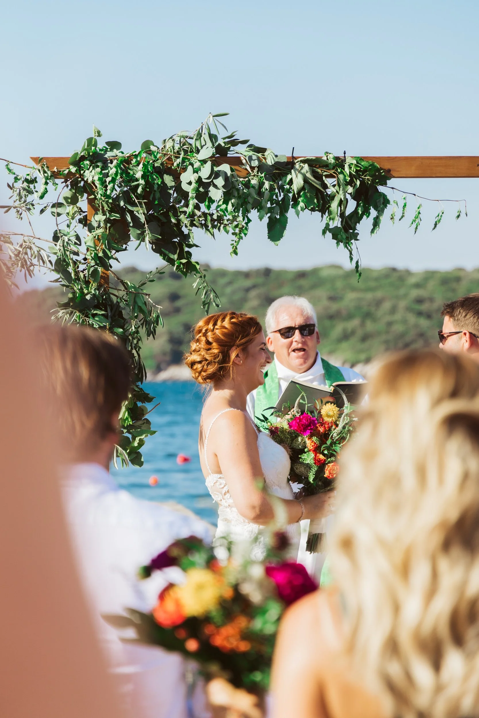 A beach wedding ceremony with a bride, groom, and officiant under a wooden arch decorated with greenery and flowers on a sunny day.