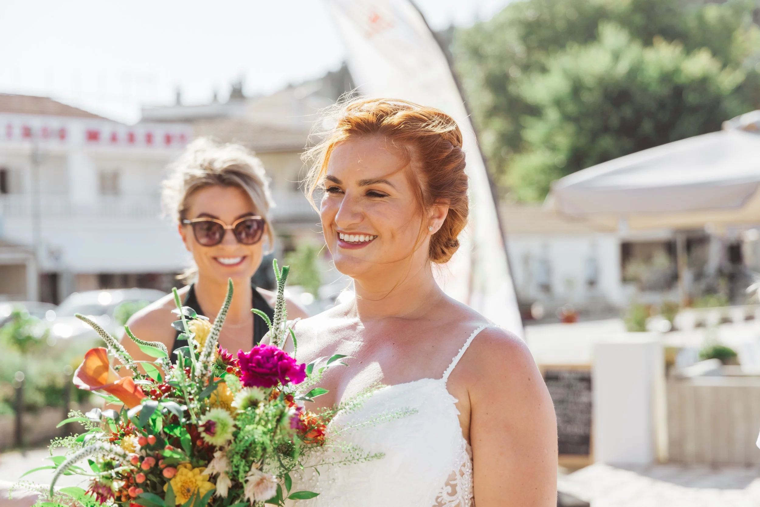 A bride with red hair holding a colorful bouquet, smiling outdoors with a woman in sunglasses in the background.