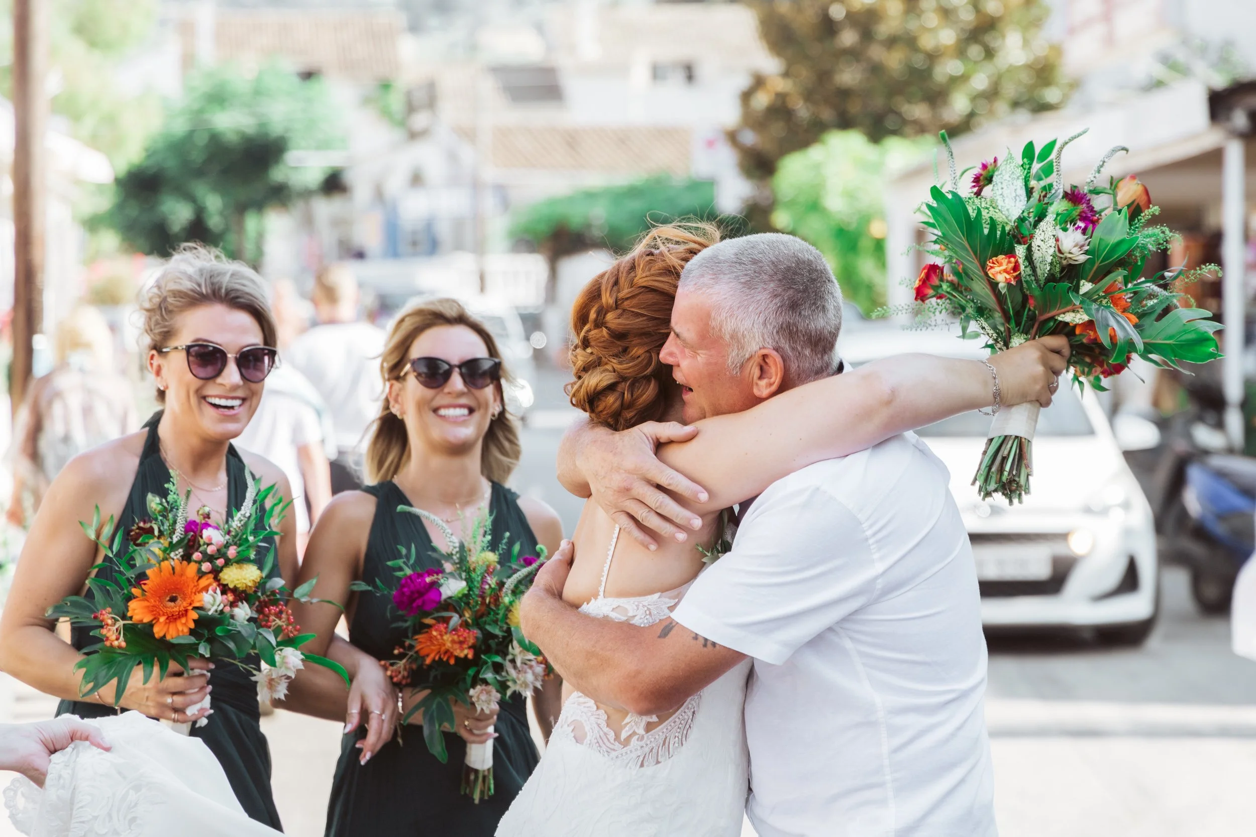 A bride and an older man hugging as they celebrate a wedding, with two bridesmaids smiling and holding bouquets, outdoors on a sunny day.
