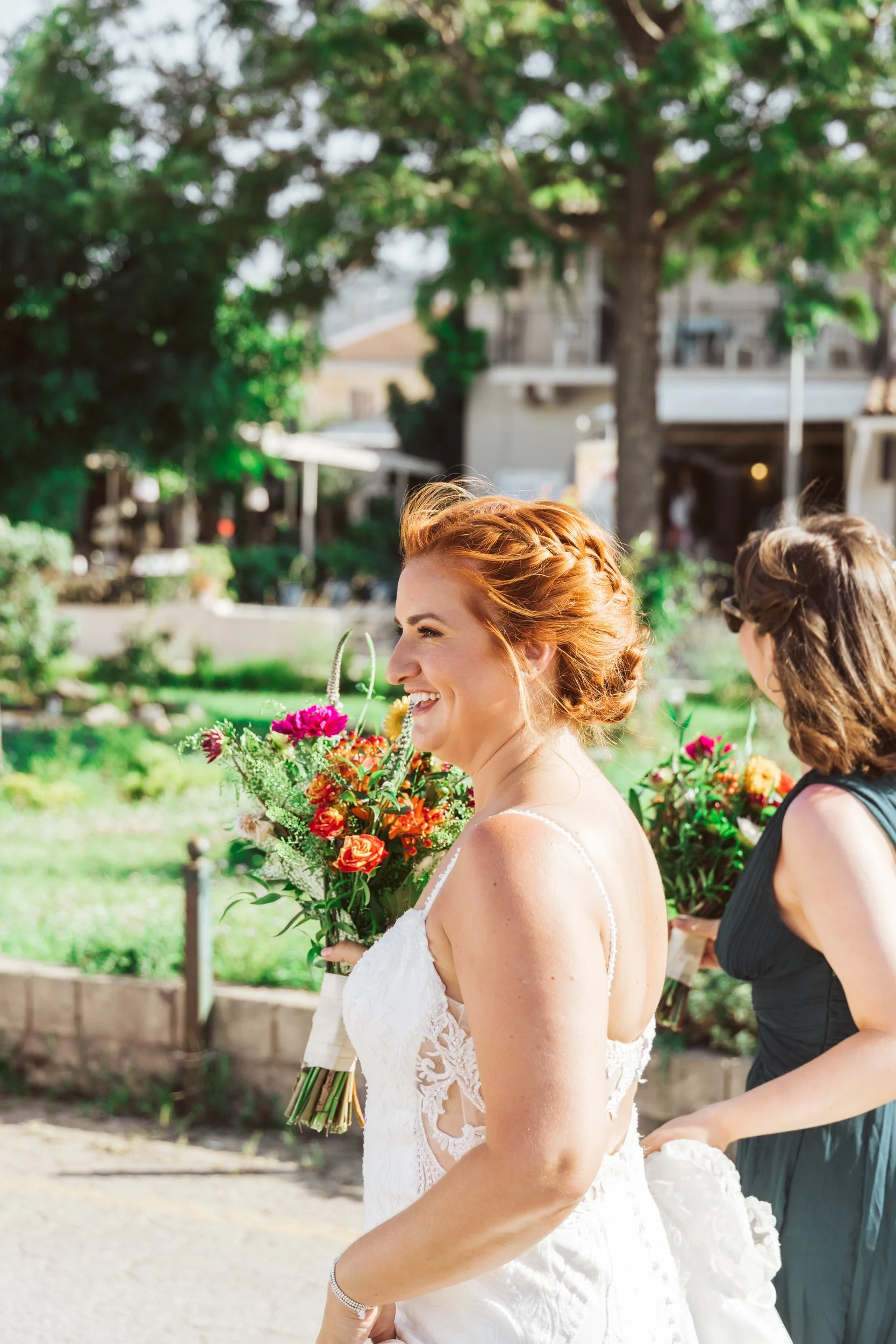 A smiling woman in a white lace wedding dress holding a colorful bouquet of flowers in an outdoor park or garden setting with greenery and trees in the background.