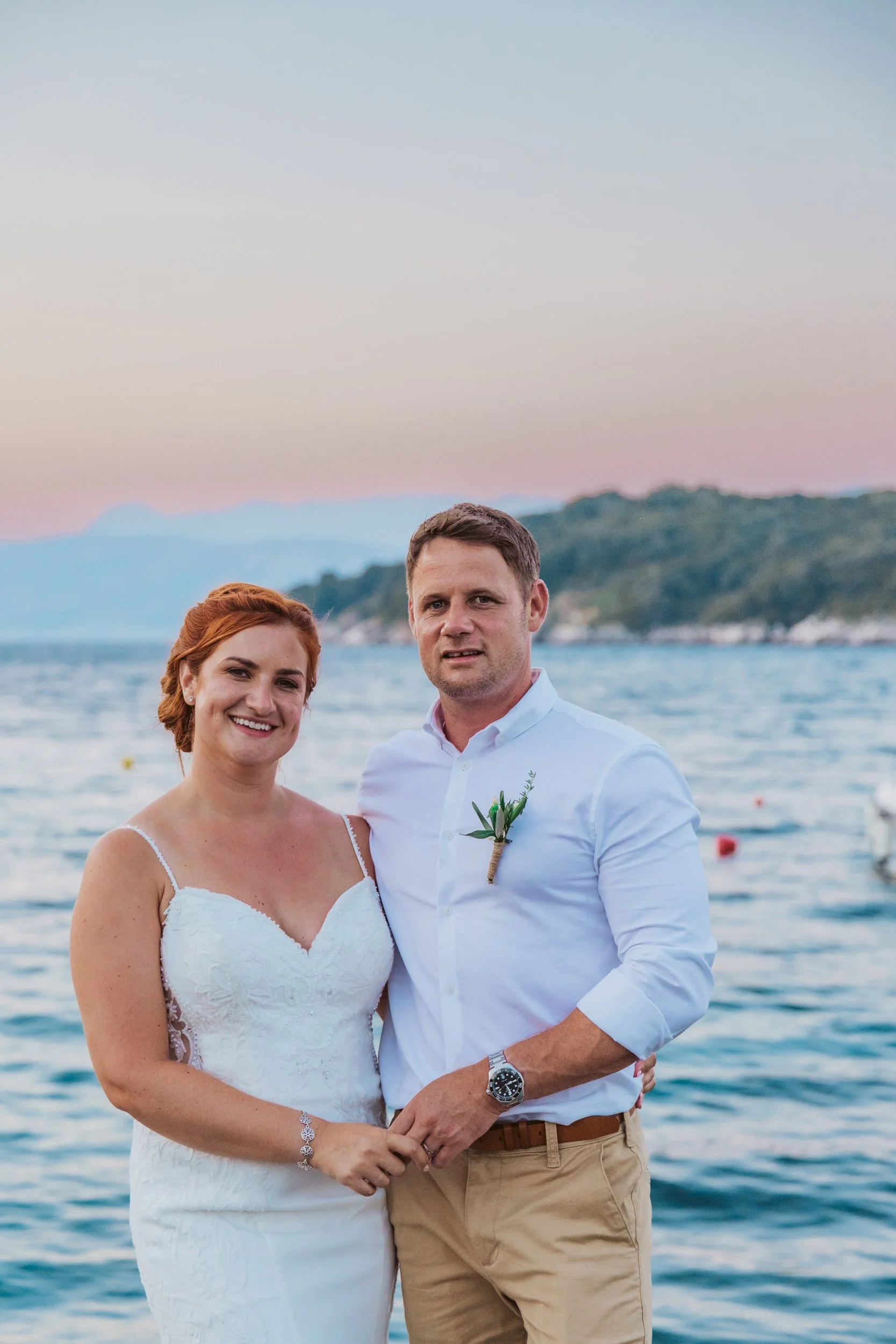 A newlywed couple at the beach during sunset, standing close together and smiling, with water and distant hills in the background.