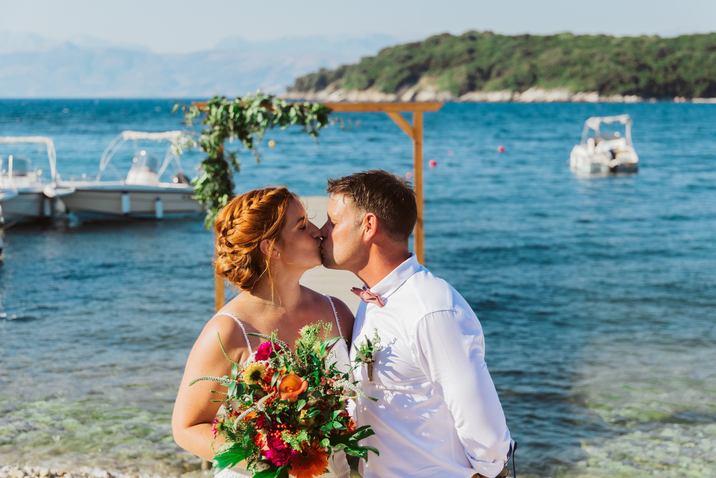 A bride and groom kissing by a seaside during their wedding, with boats and a mountainous landscape in the background.