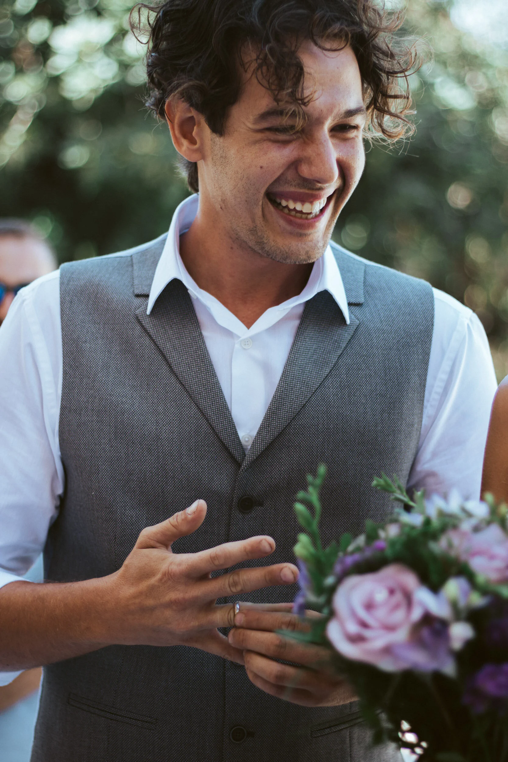A man with curly dark hair, dressed in a white shirt and gray vest, smiling and holding a bouquet of pink and purple flowers during an outdoor event.