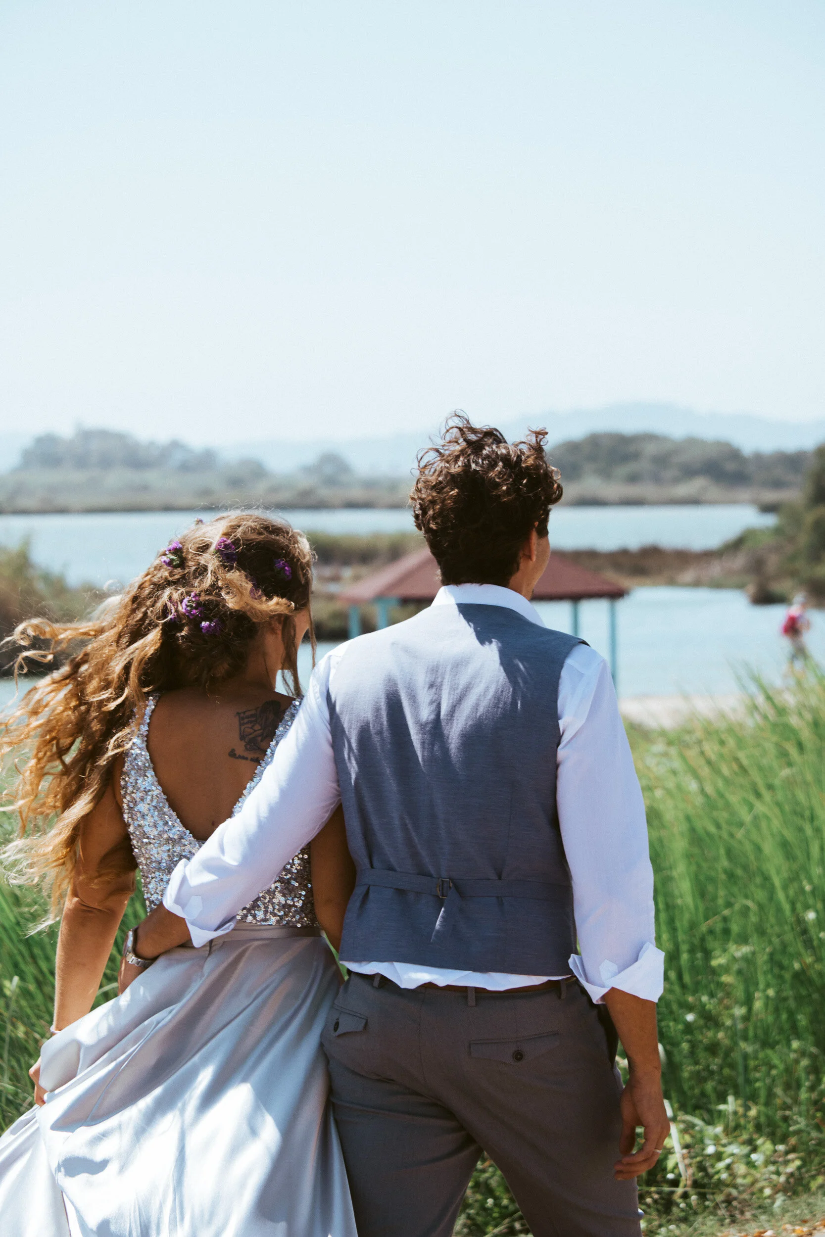 A couple walking near a water body, the woman wearing a sparkly dress and the man dressed in a vest and shirt, with lush green surroundings and a gazebo in the background.