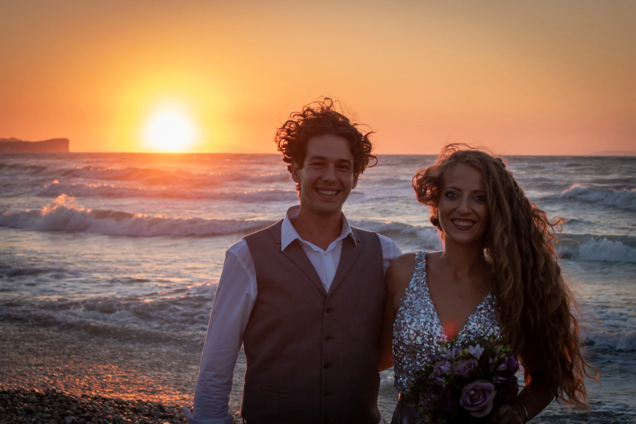 A smiling man and woman stand together on a beach at sunset, with waves in the background. The woman holds a bouquet of flowers and they are dressed in formal attire.