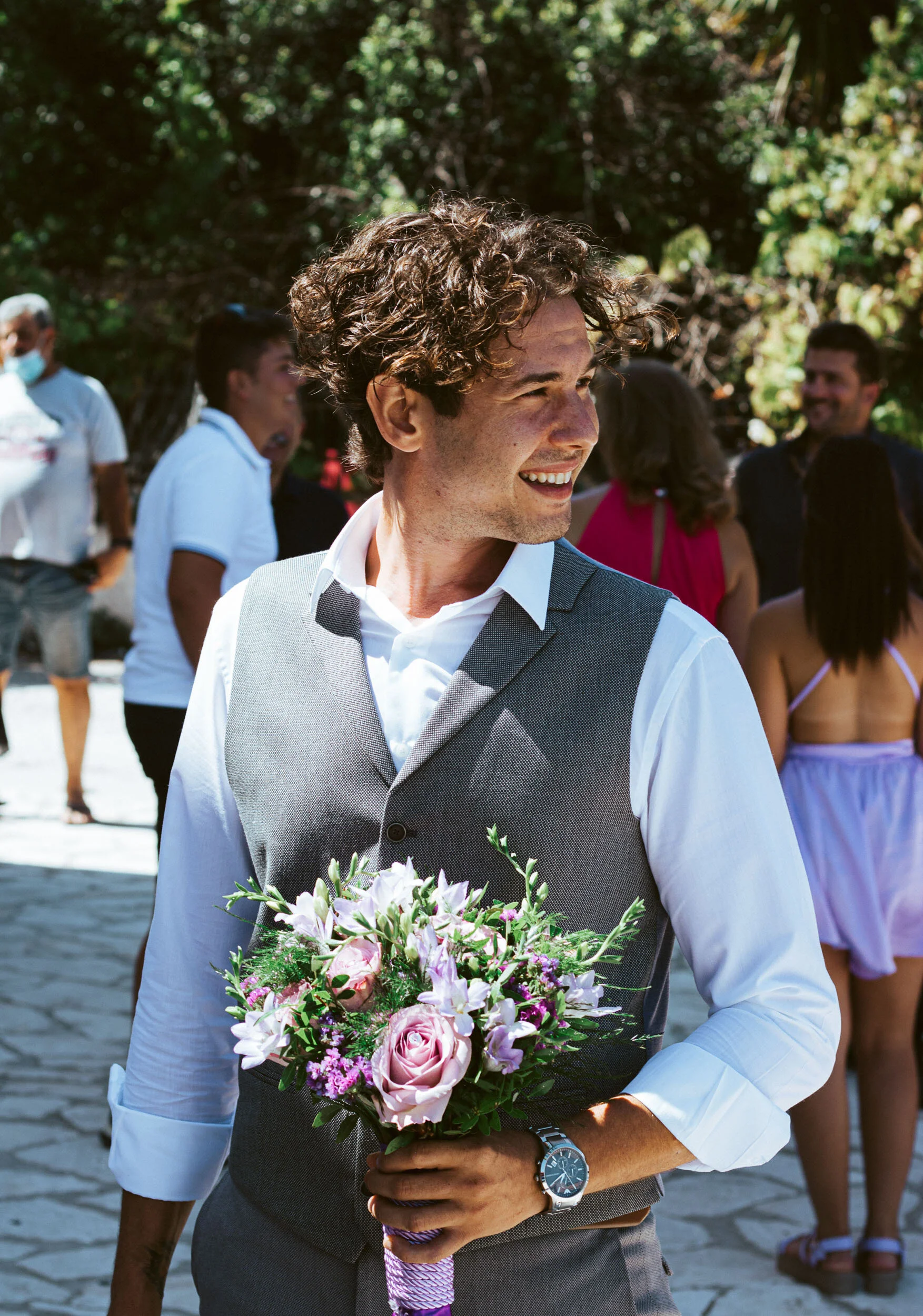 A man with curly hair smiling and holding a bouquet of pink and white flowers during a celebration outdoors, with several people in the background.