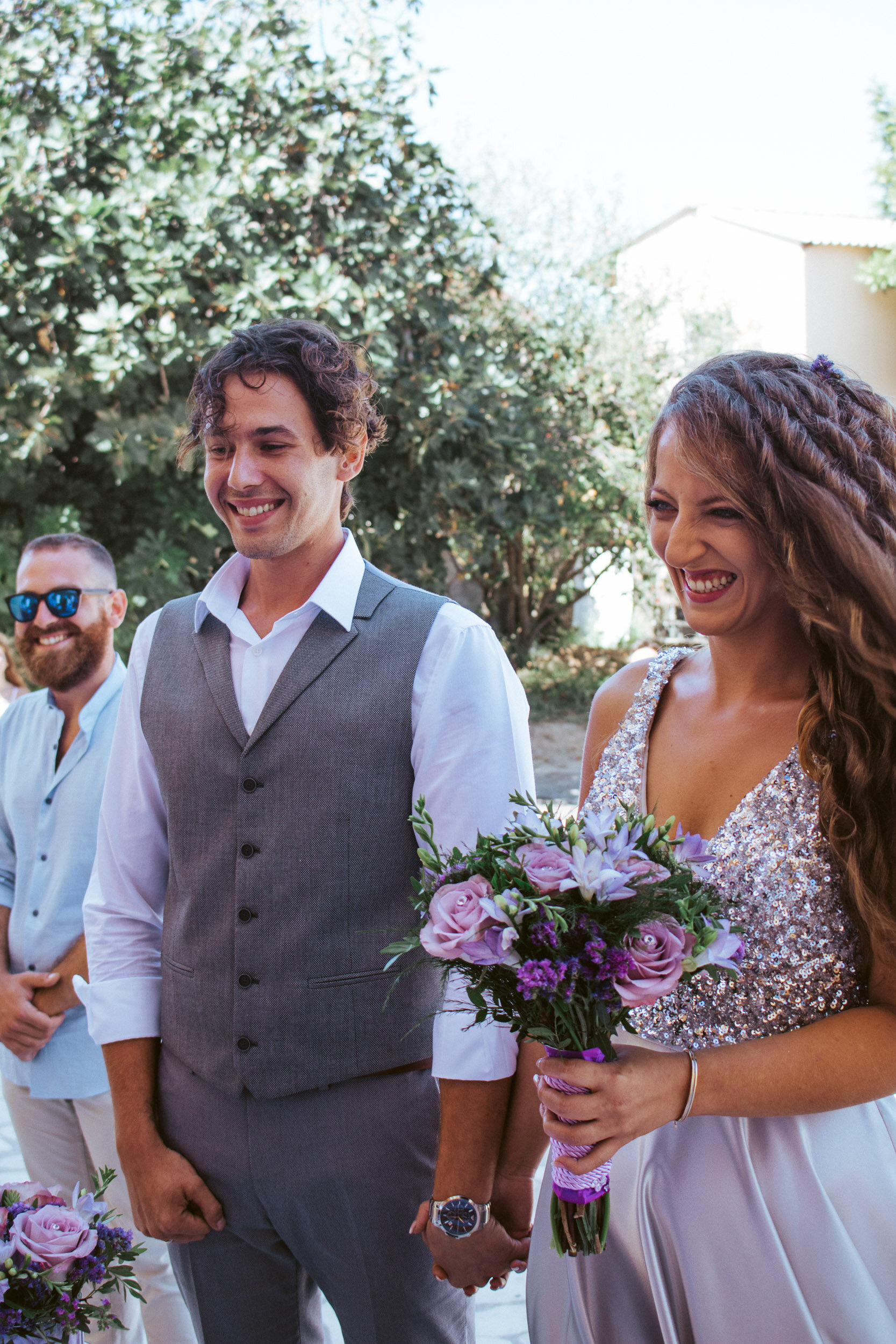 A bride and groom holding hands at their wedding outside, with friends in the background, surrounded by trees and greenery.
