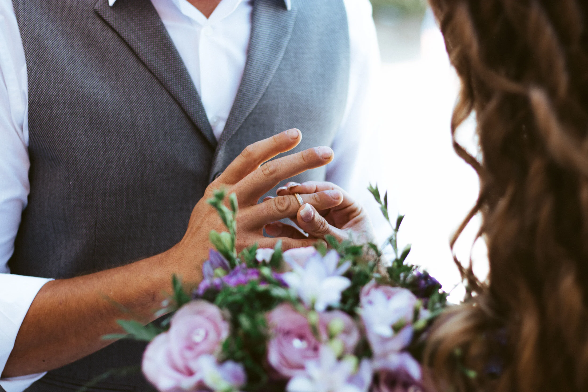 A person wearing a gray vest and white shirt is holding a ring to another person's finger during a wedding ceremony, with a bouquet of purple, pink, and white flowers in the foreground.