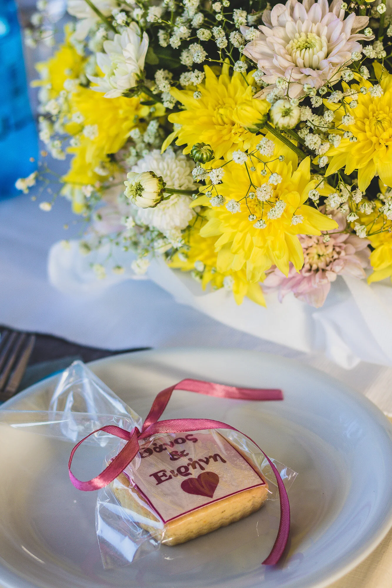 A gift wrapped cookie with a pink ribbon and a heart and text, placed on a white plate, with a bouquet of yellow, white, and pink flowers in the background.