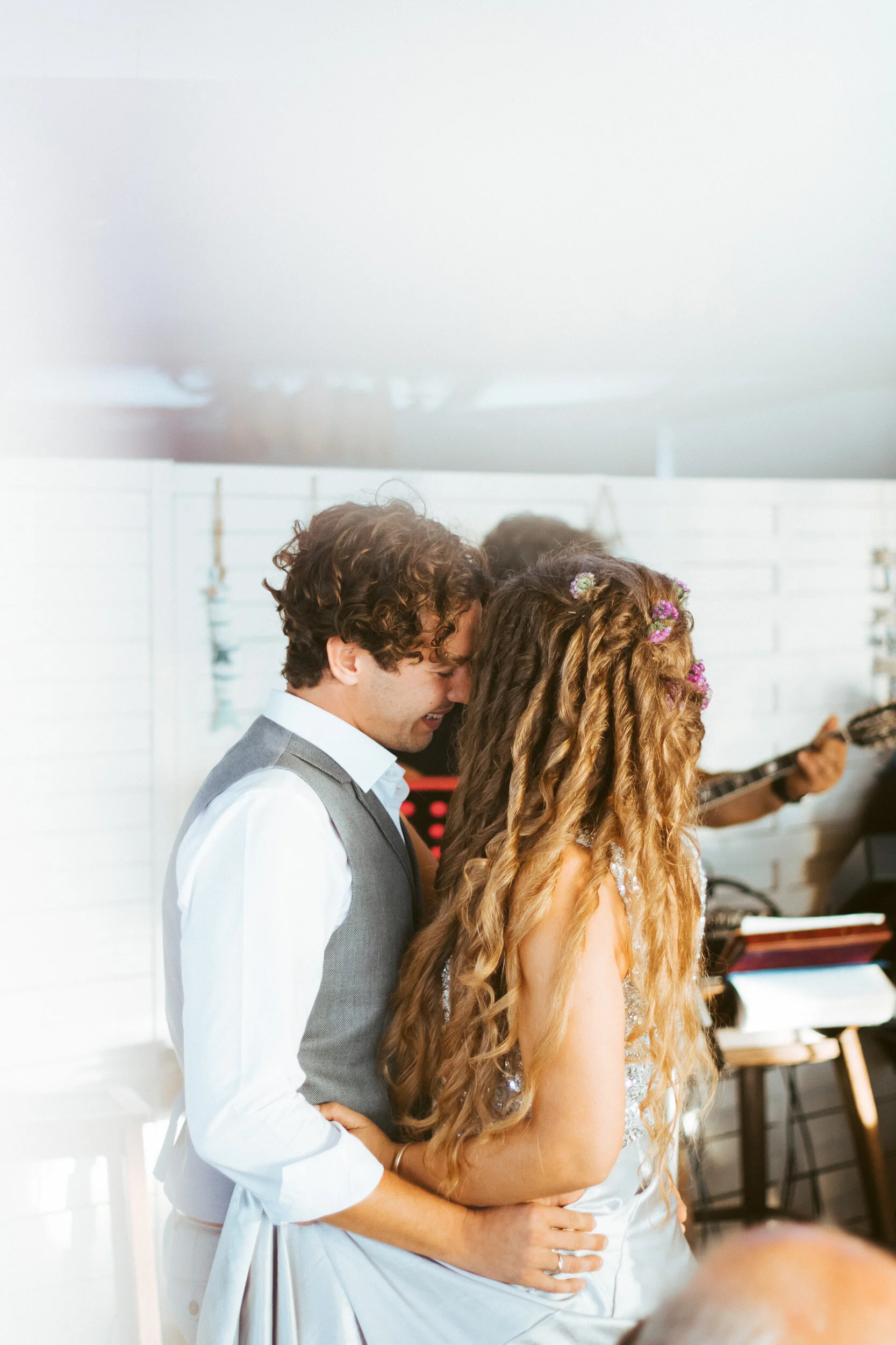 A couple sharing a close moment, possibly a wedding, with a man in a gray vest and a white shirt and a woman with long, wavy hair with flowers, smiling and embracing.