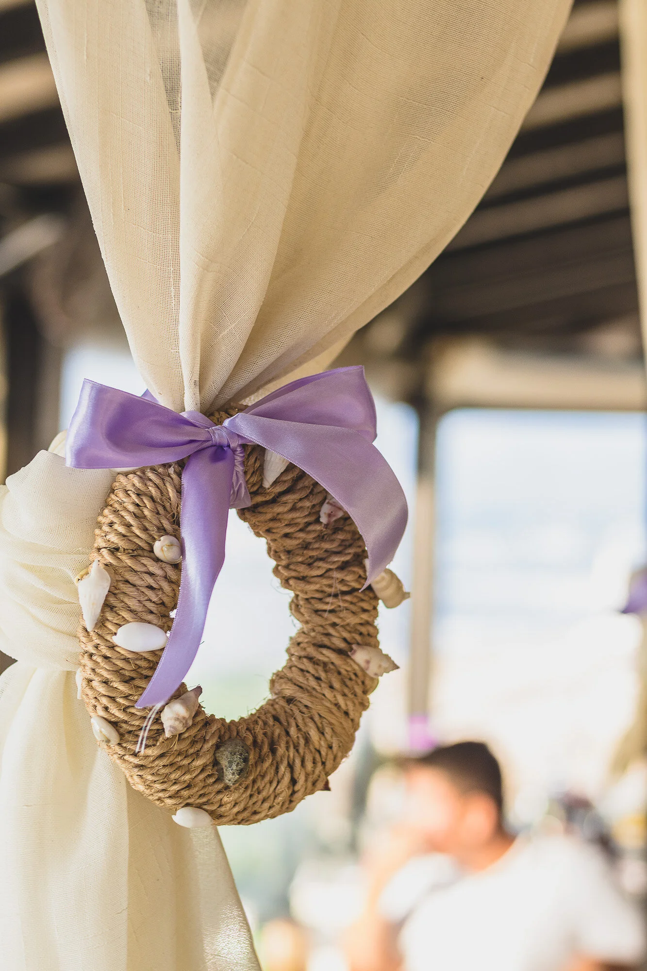 A decorative wreath made of thick twine with seashells attached, tied with a lavender ribbon, hanging on a beige curtain.