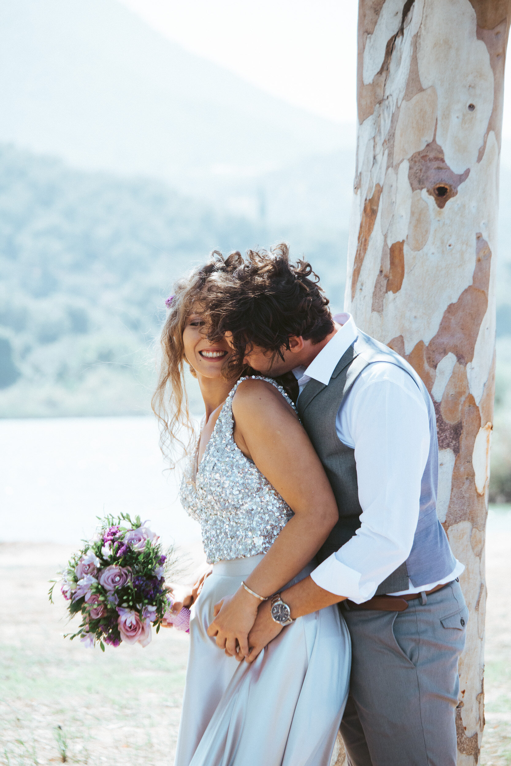 A happy couple embrace outdoors during a sunny day. The woman holds a bouquet and wears a sparkly top and long skirt, while the man wears a vest and dress shirt.