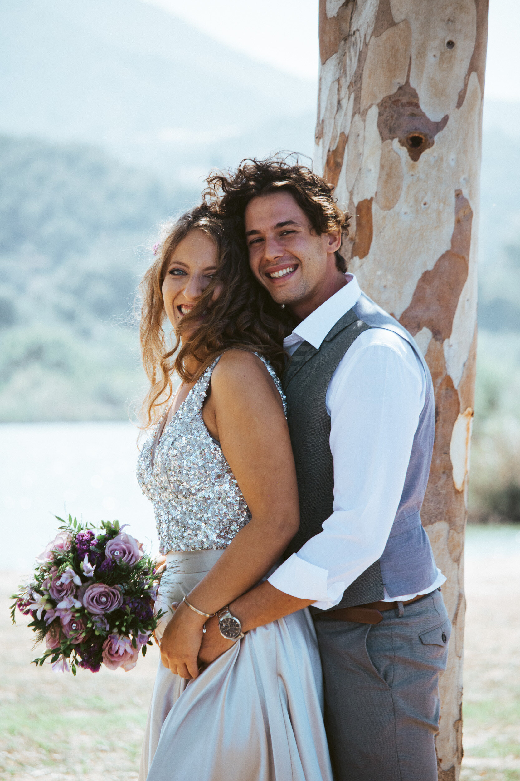 A newlywed couple standing outdoors during daytime, smiling and embracing near a tree with a blurred background of mountains and sky.