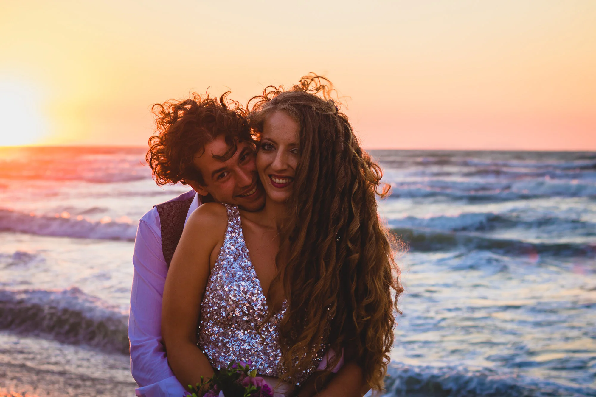 A happy couple standing on the beach during sunset, with the ocean waves in the background. The woman has long, curly hair and is wearing a sparkly dress. The man has curly hair and is wearing a light-colored shirt with a vest. They are smiling and e