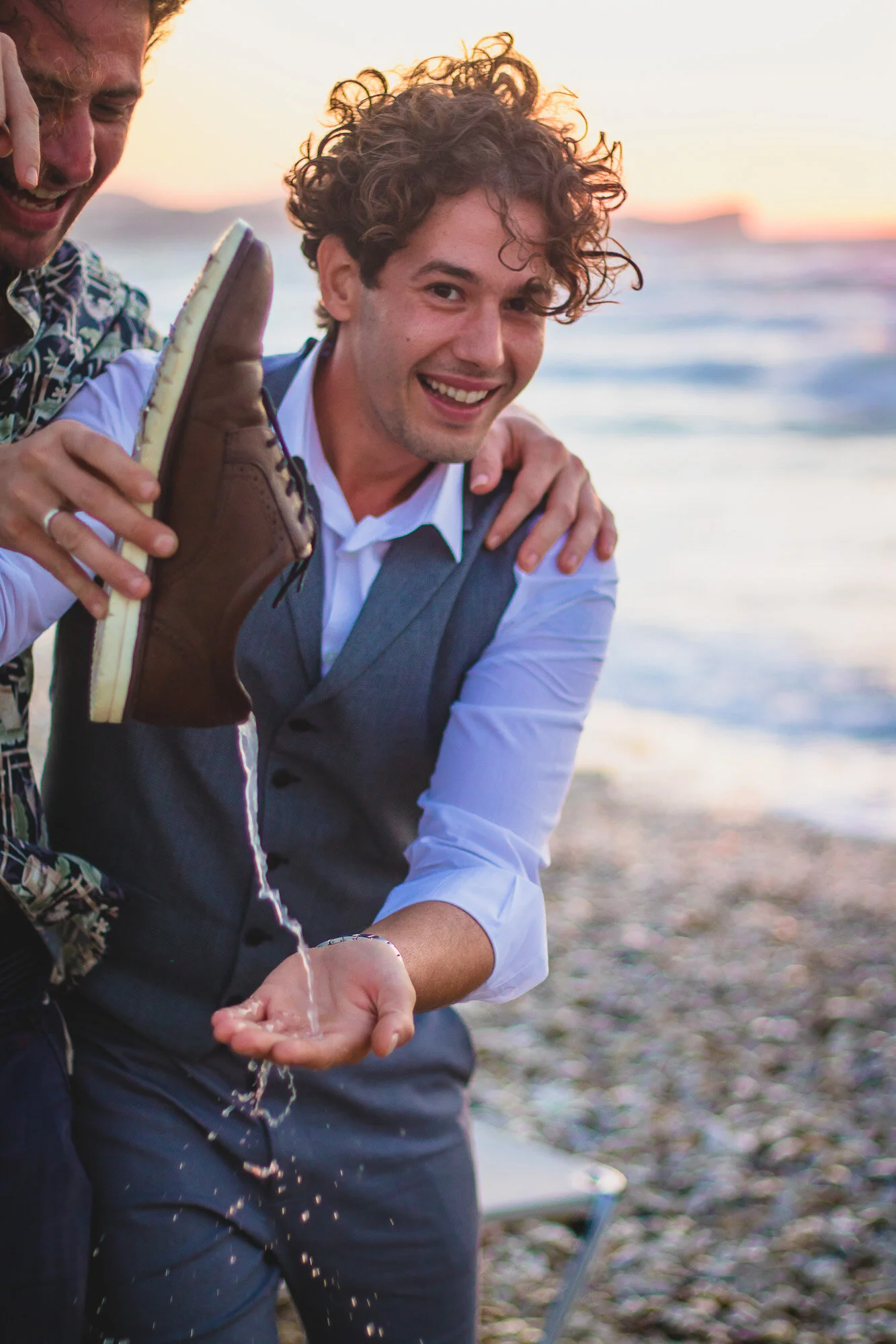 Two men at the beach, one holding a shoe and pouring water into his hand, smiling and enjoying a sunset.
