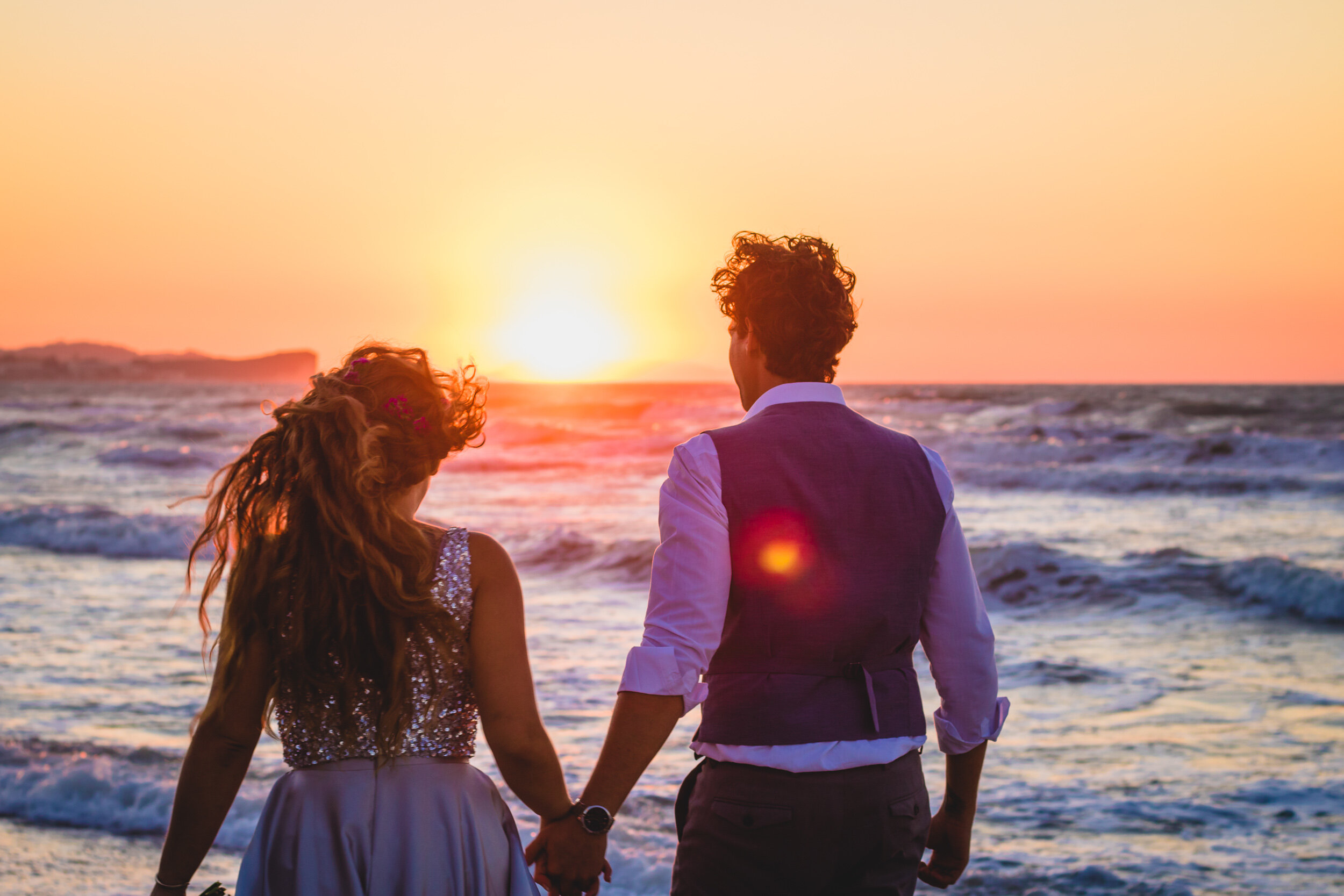 A couple holding hands at sunset on the beach, facing the ocean with the sun setting on the horizon.