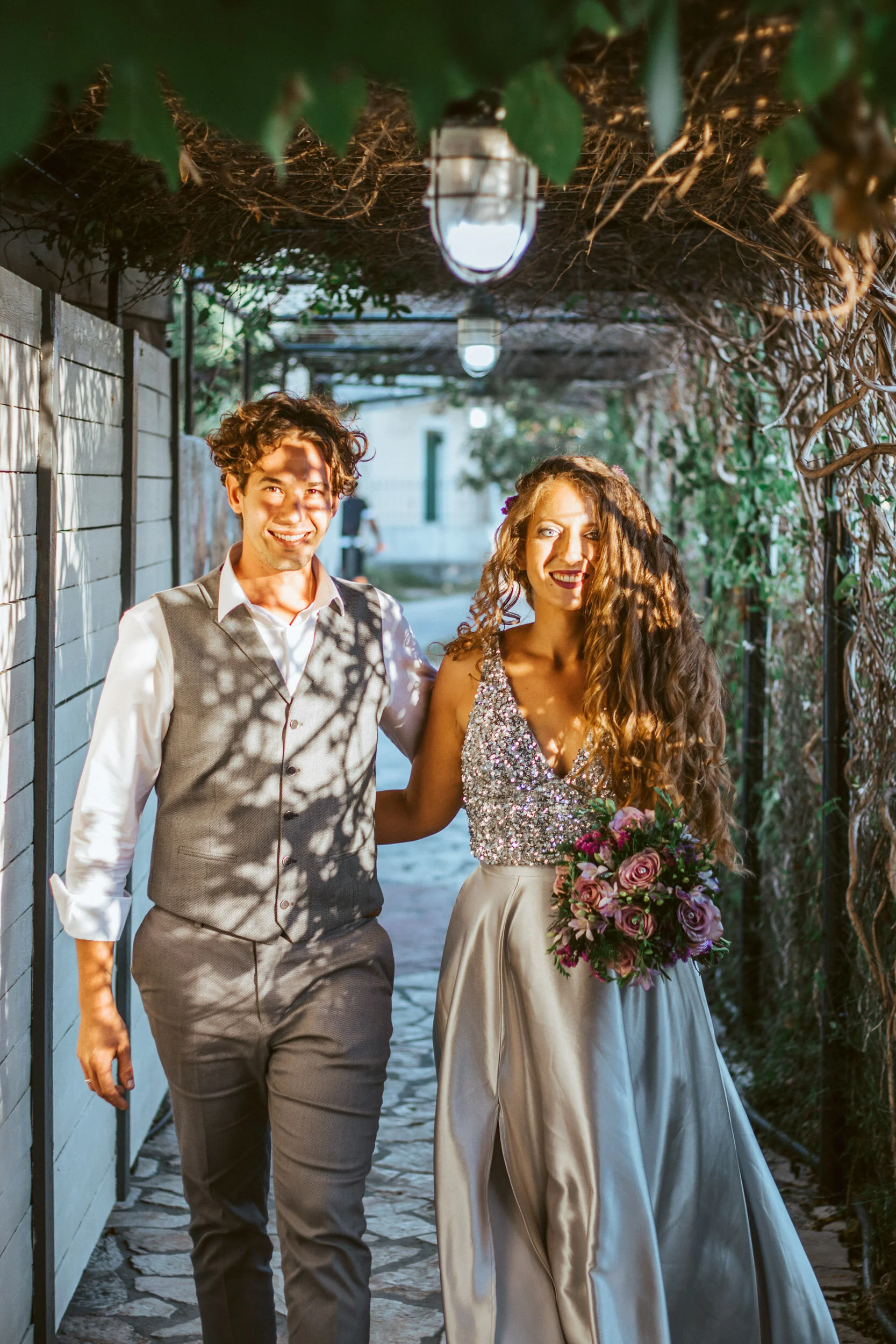 A couple walking together in an outdoor setting, with trees and hanging lights overhead. The woman is wearing a shiny, sleeveless dress and holding a bouquet of flowers; the man is dressed in a light-colored vest and shirt.