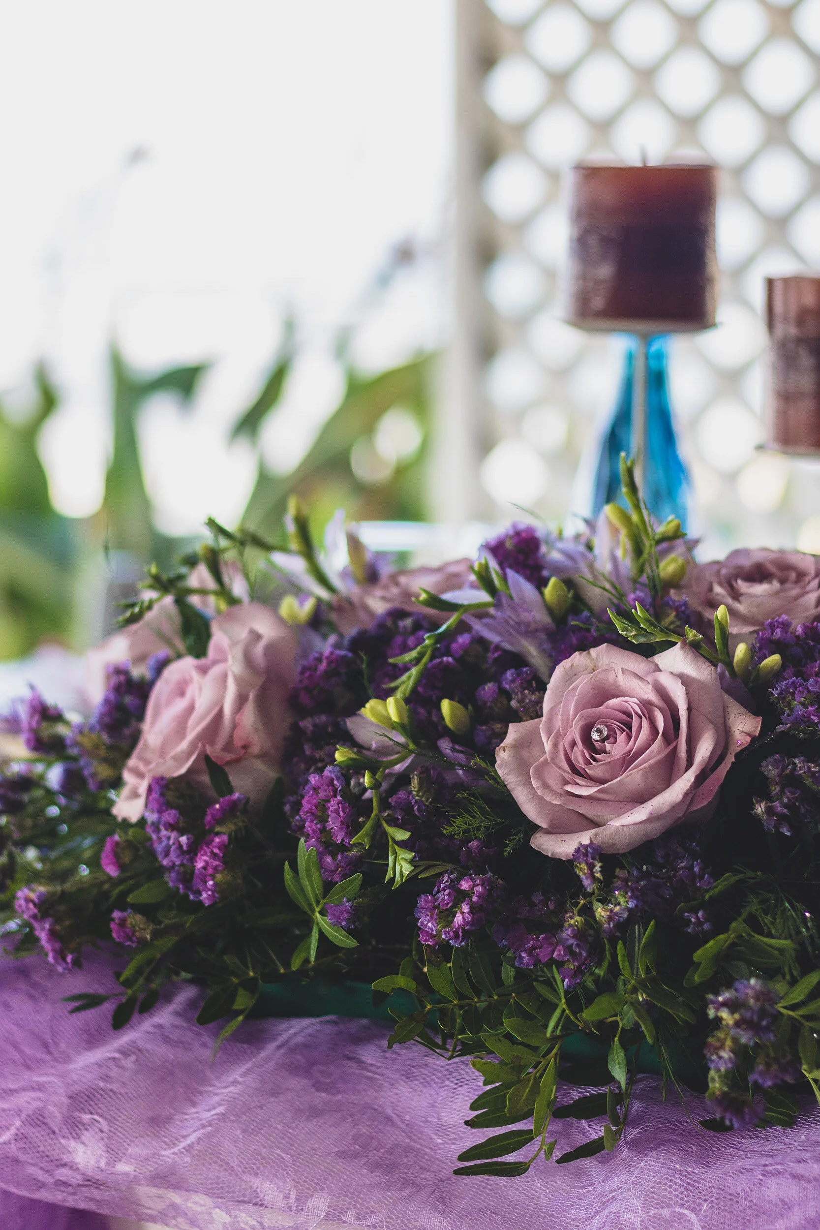 Close-up of a purple and pink floral arrangement with roses and greenery, on a pink tablecloth, with blurred background.