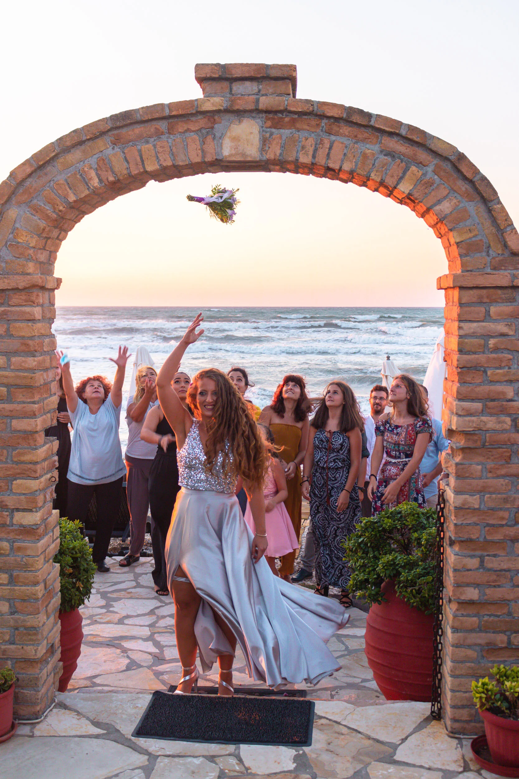 A woman in a shiny silver dress throws a bouquet of flowers into the air at a beachside wedding, with guests watching and smiling in the background, ocean waves in the distance.