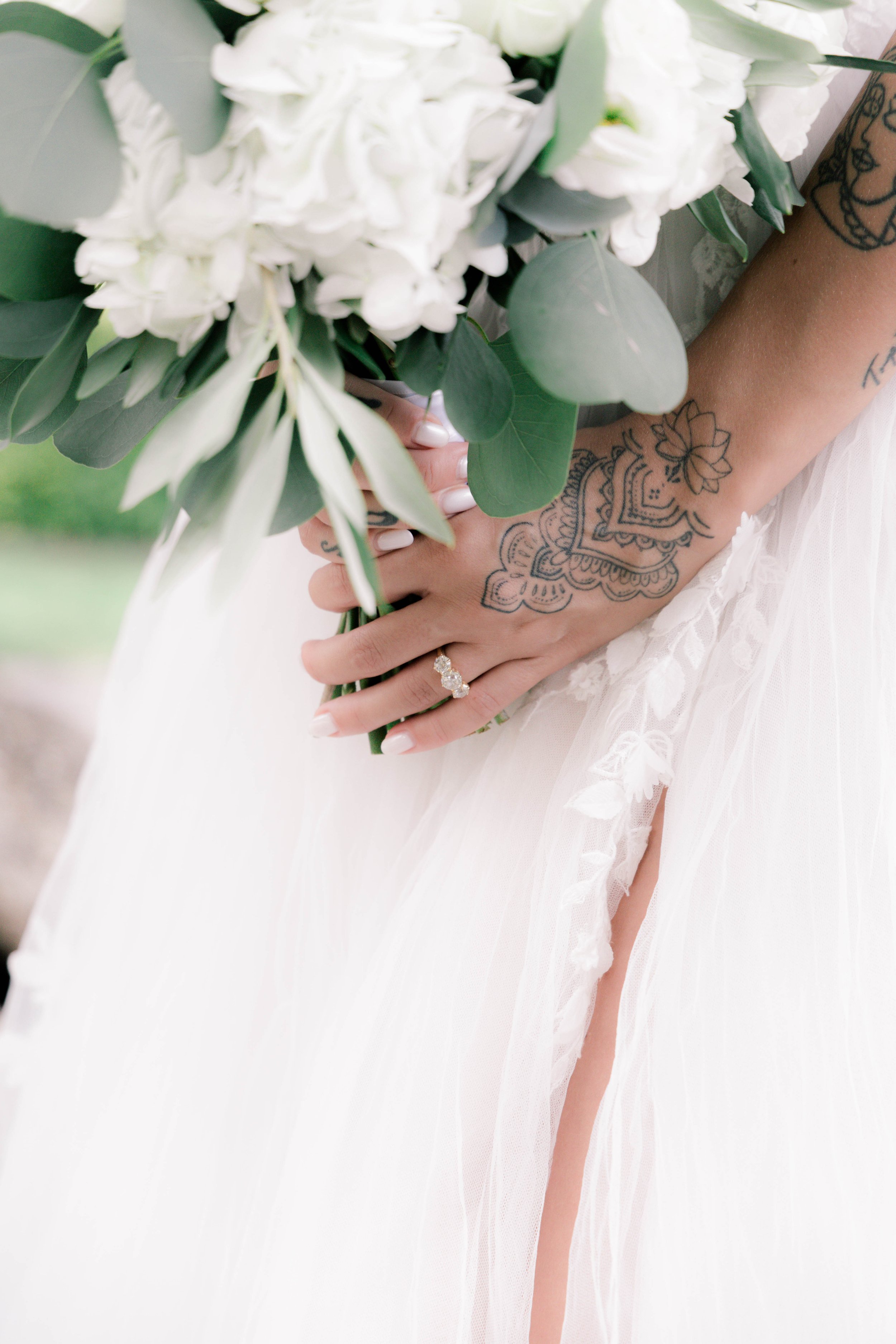 A bride holding a bouquet of white flowers and green leaves, showing her hand with an engagement ring and a colorful tattoo, wearing a white wedding dress.