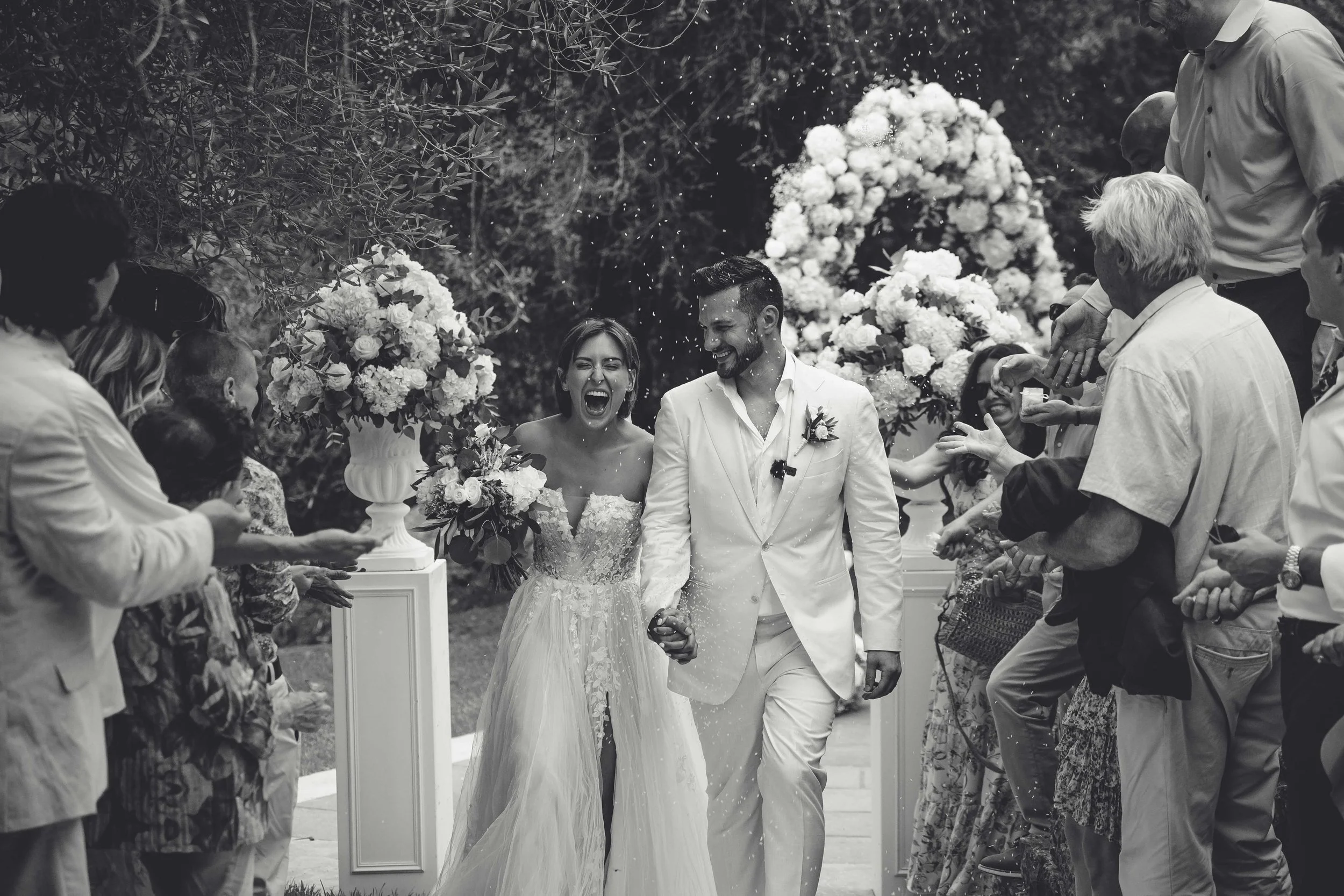 A black and white photograph of a bride and groom at their wedding, walking hand in hand through an outdoor aisle lined with guests on both sides, who are throwing confetti in celebration.
