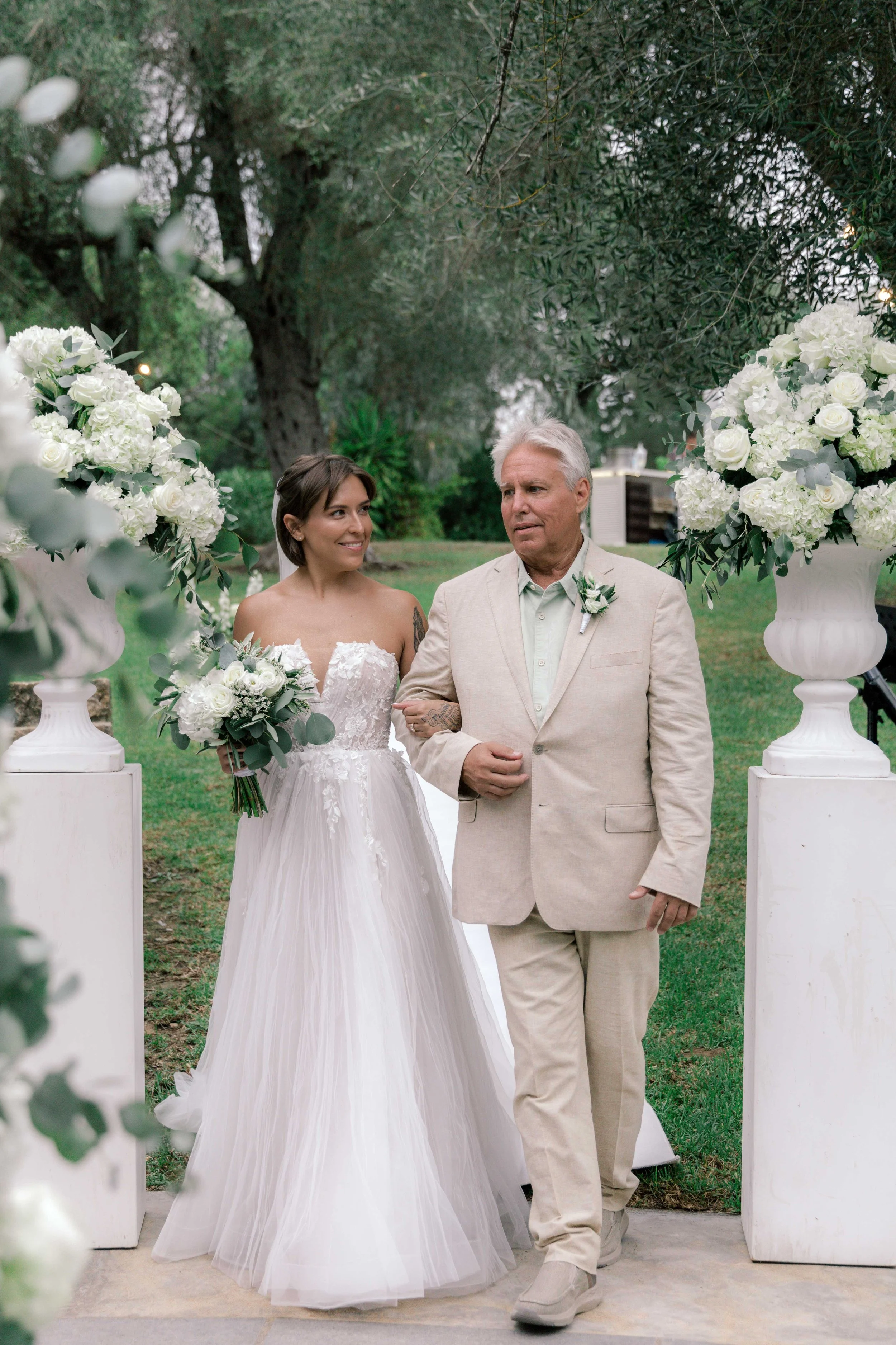 Bride walking down the aisle with her father at an outdoor wedding ceremony, surrounded by white floral arrangements and greenery.