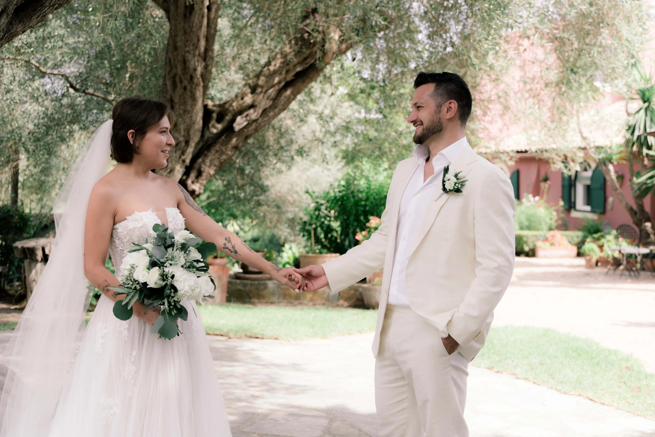 A bride and groom holding hands and smiling at each other outdoors in a garden setting during their wedding.