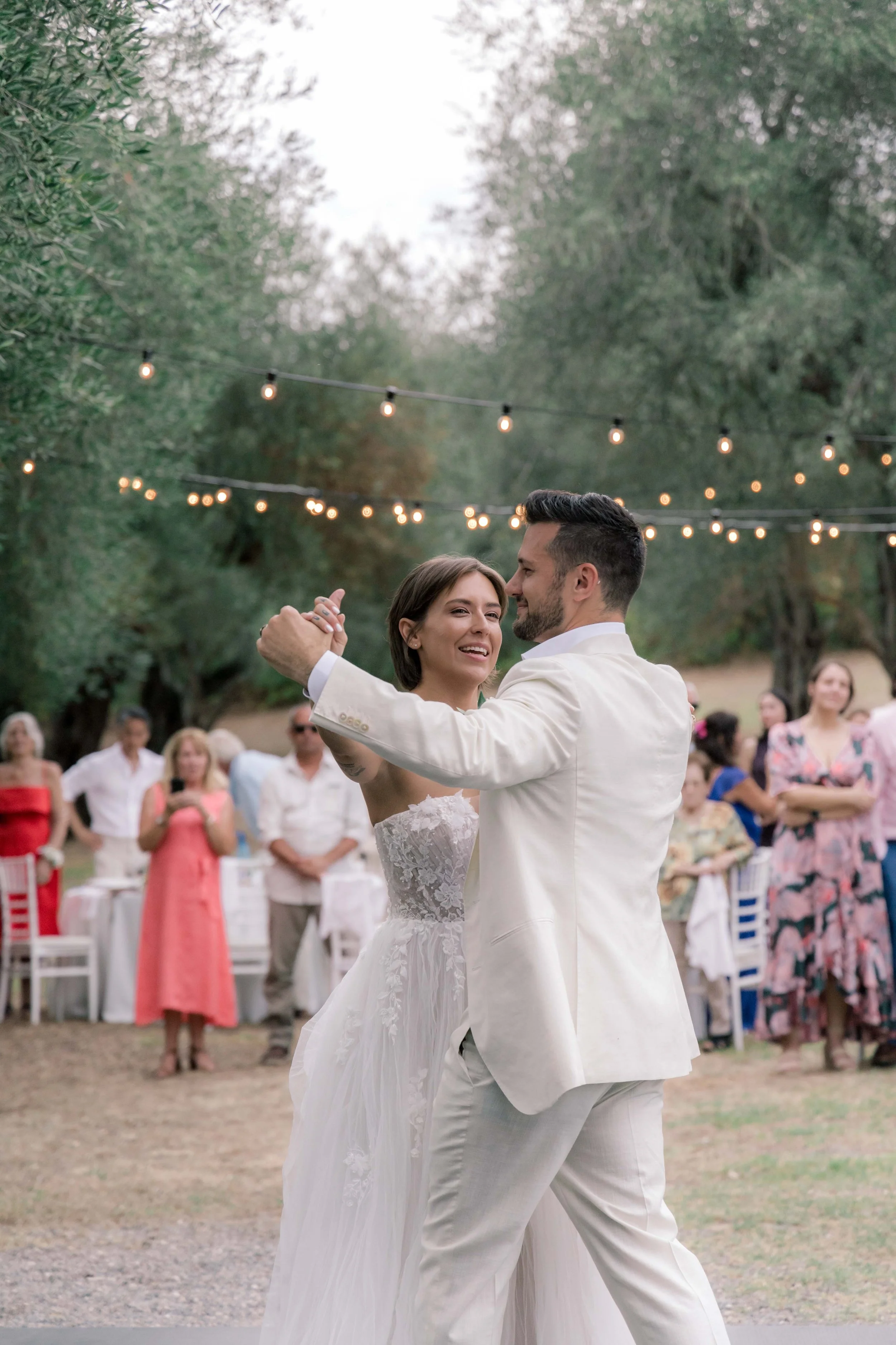 A newlywed couple dancing outdoors under string lights, with guests watching in the background.