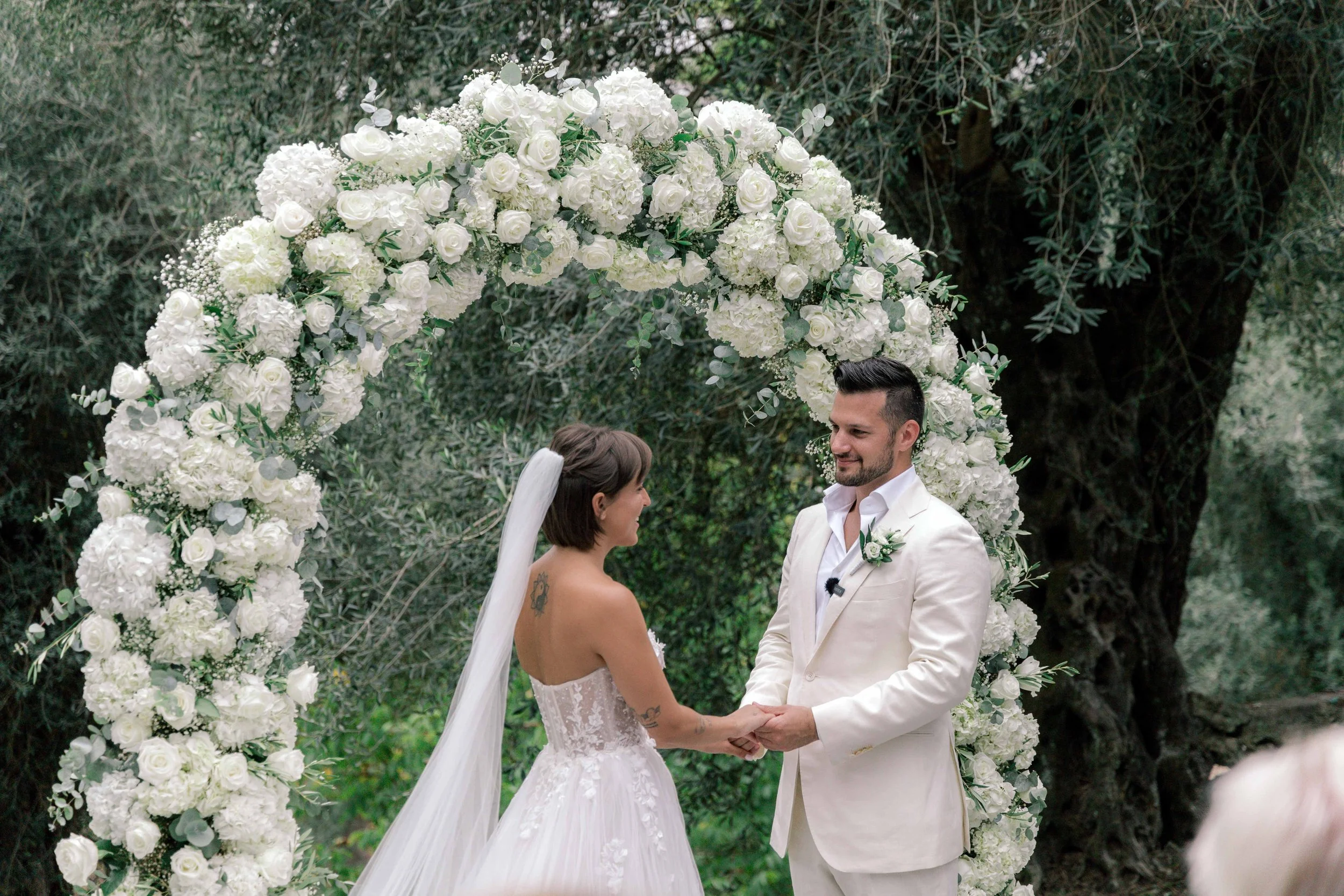 Bride and groom holding hands and facing each other under a floral arch during their wedding ceremony outdoors.
