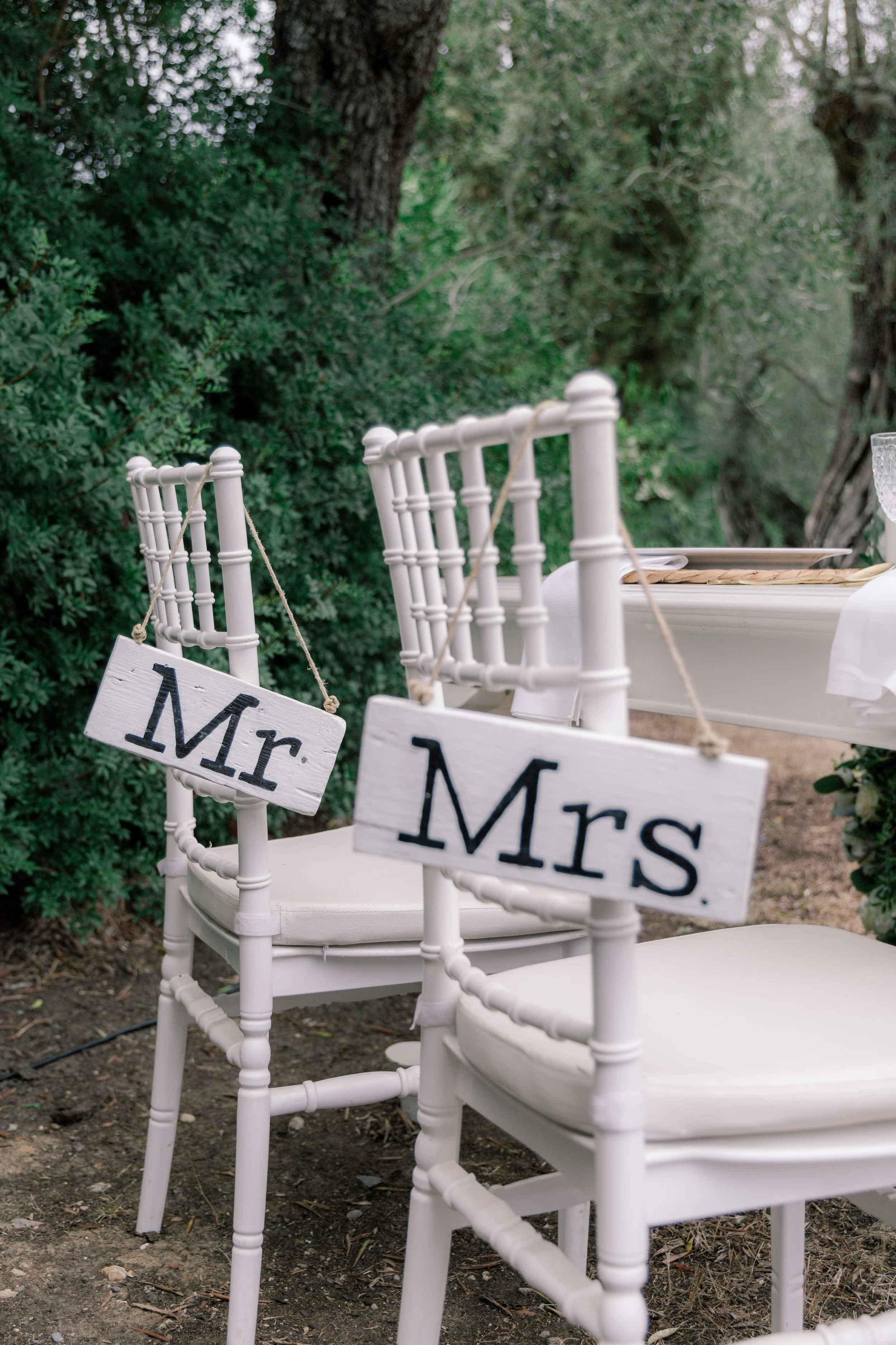 White chairs with 'Mr' and 'Mrs' signs hanging on the back, set outdoors with greenery in the background