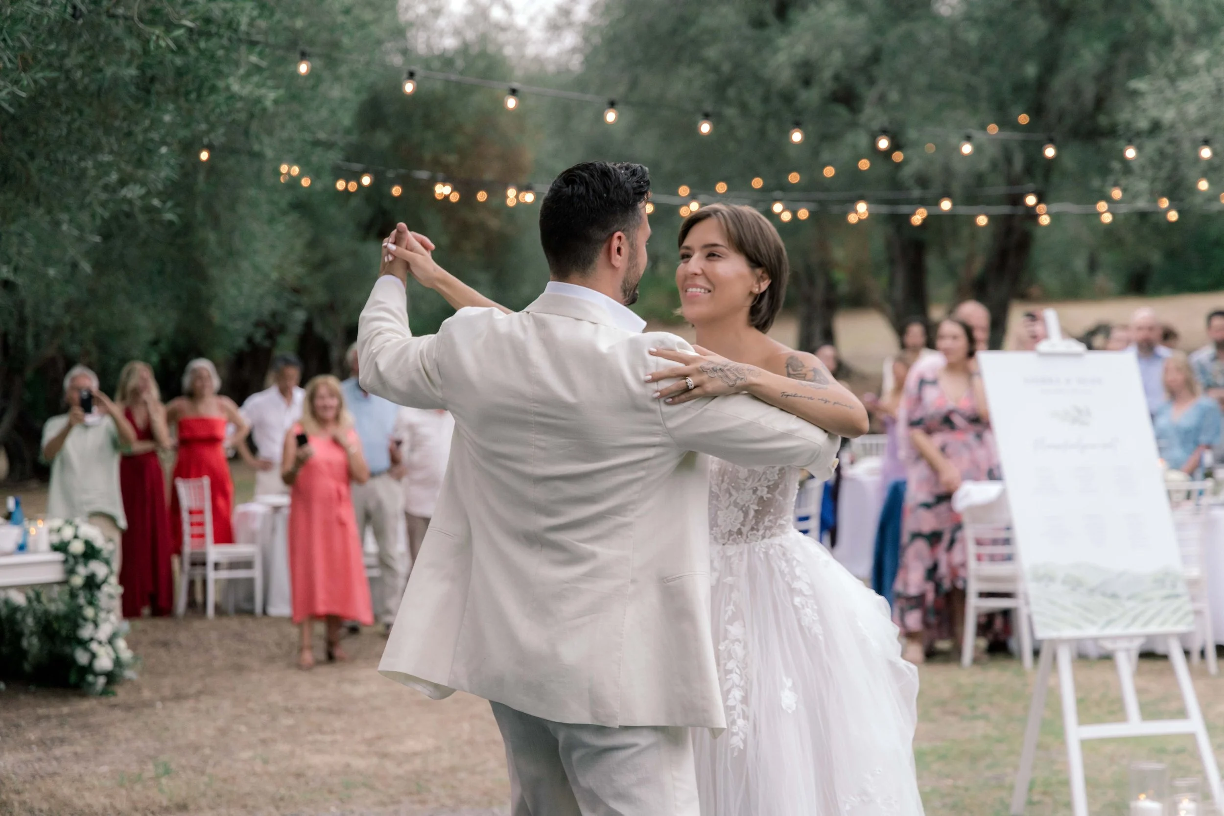 Couple dancing during wedding reception outdoors, surrounded by guests and string lights hanging above.