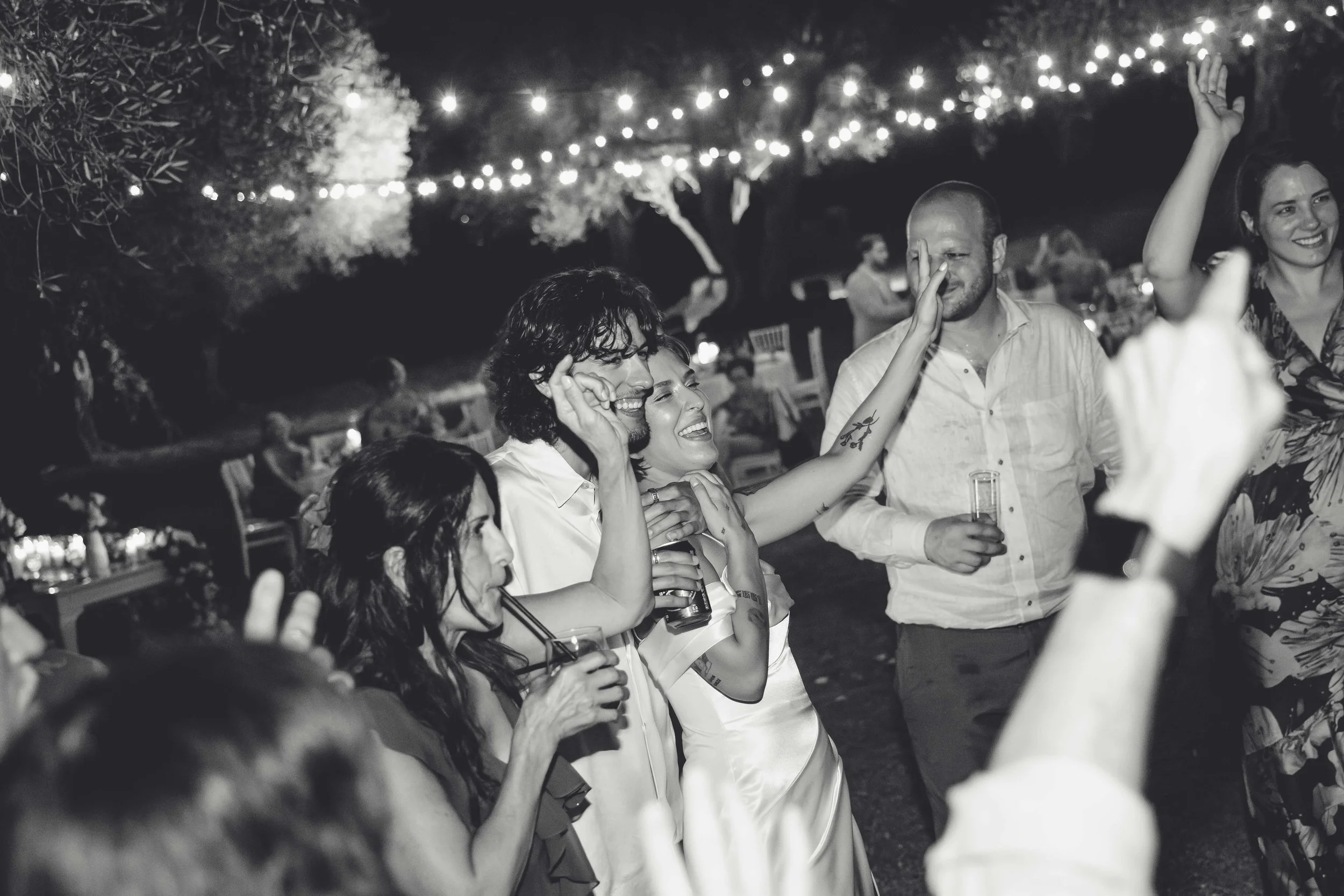 People celebrating at a nighttime outdoor event with string lights overhead. Several people are smiling and holding drinks, engaging in joyful conversation and dancing.