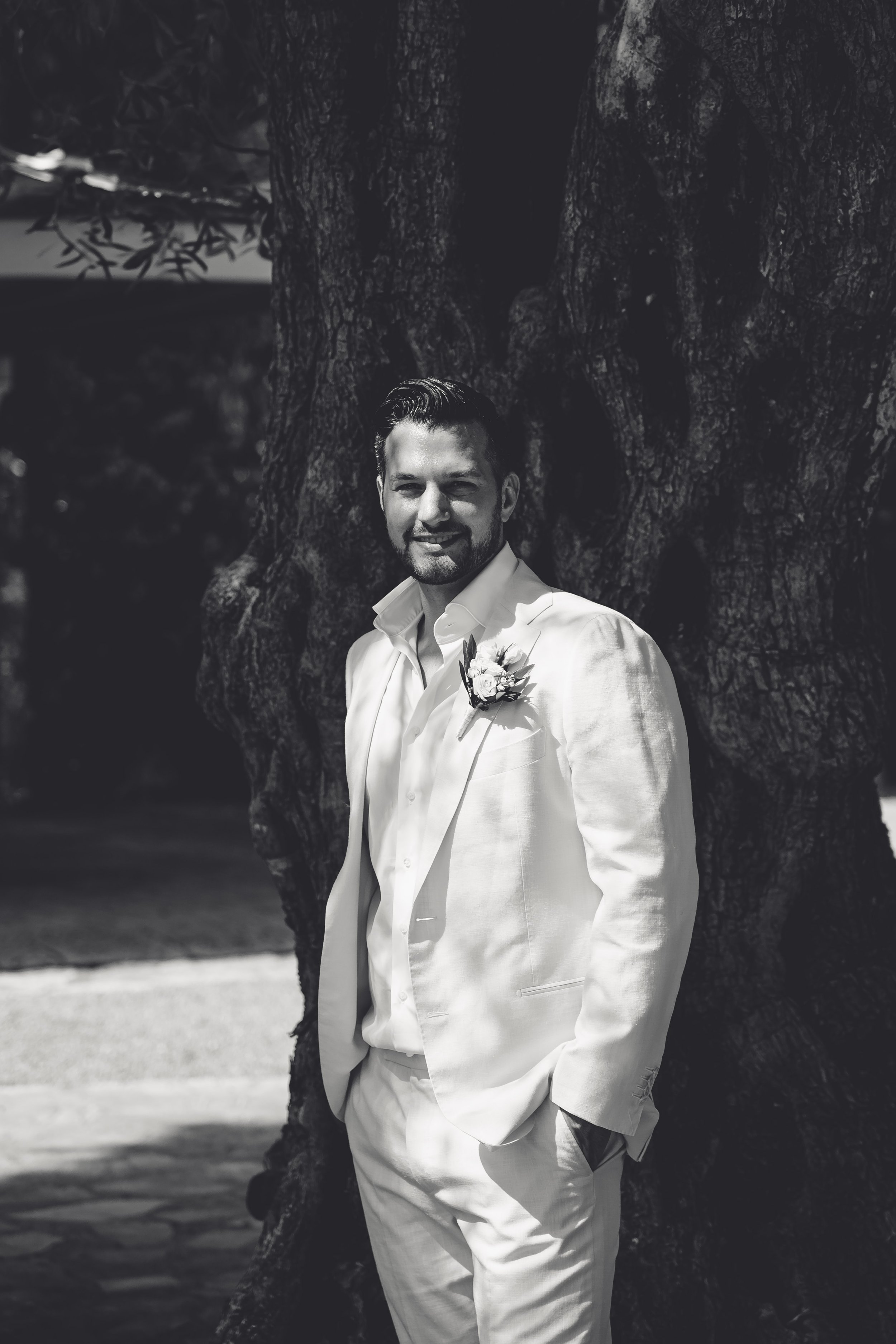 A man in a white suit with a boutonniere, smiling and leaning against a large tree outdoors.