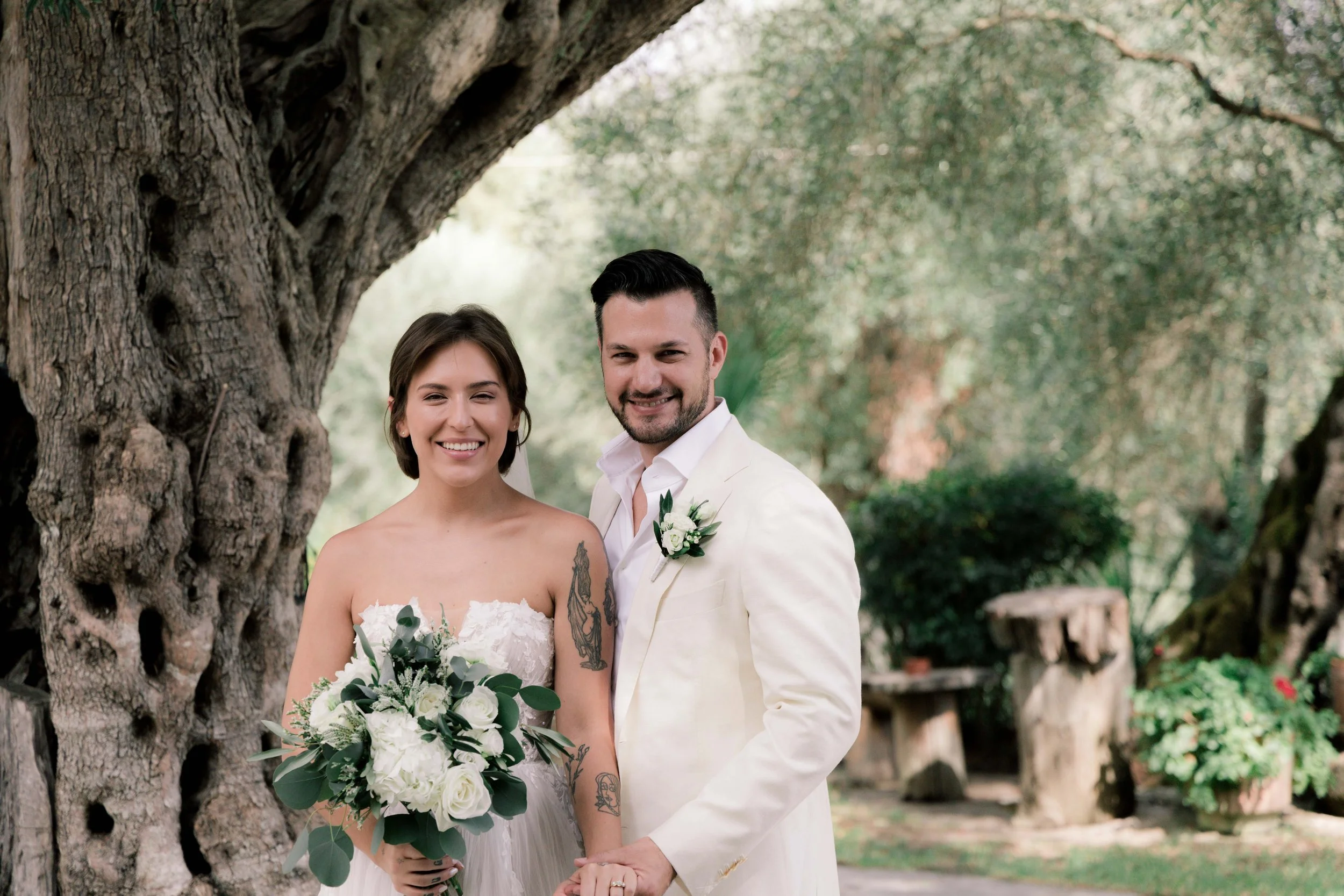 A bride and groom standing outdoors under a large tree, smiling at the camera. The bride is holding a bouquet of white flowers and wearing a strapless wedding dress. The groom is dressed in a light-colored suit with a boutonniere. The background feat