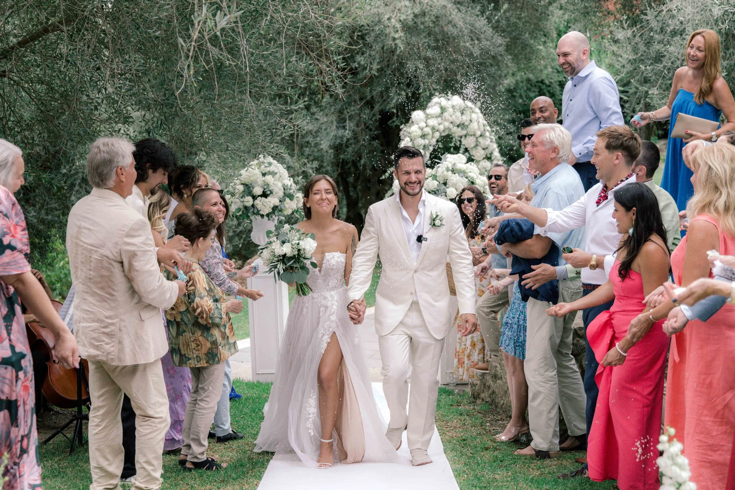 A wedding celebration with a bride in a white dress and a groom in a white suit holding hands and walking down an outdoor aisle. Guests on both sides throw flower petals and smile under a canopy of greenery and white floral arrangements.