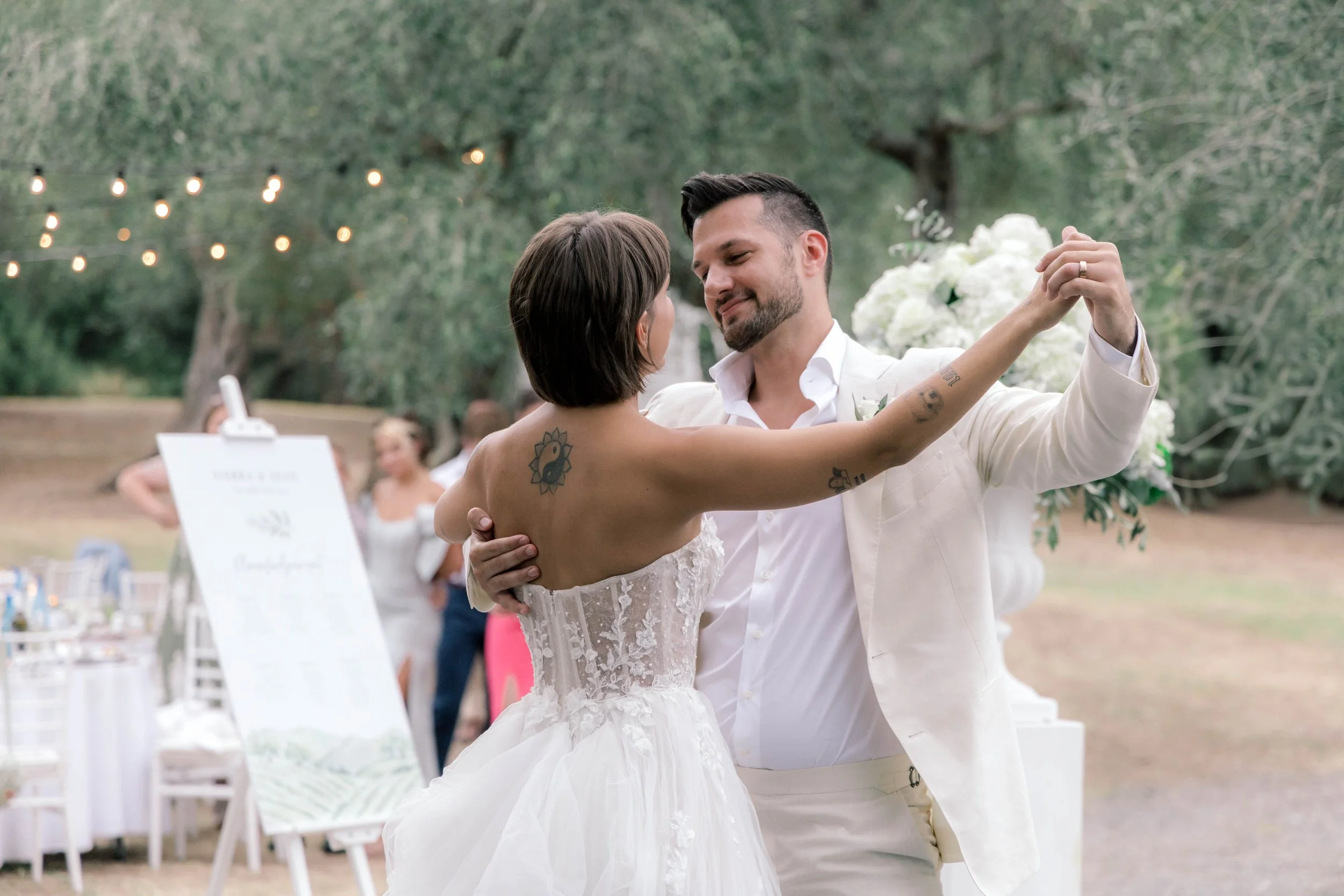 A couple dancing at an outdoor wedding reception, surrounded by string lights and trees, with guests in the background.