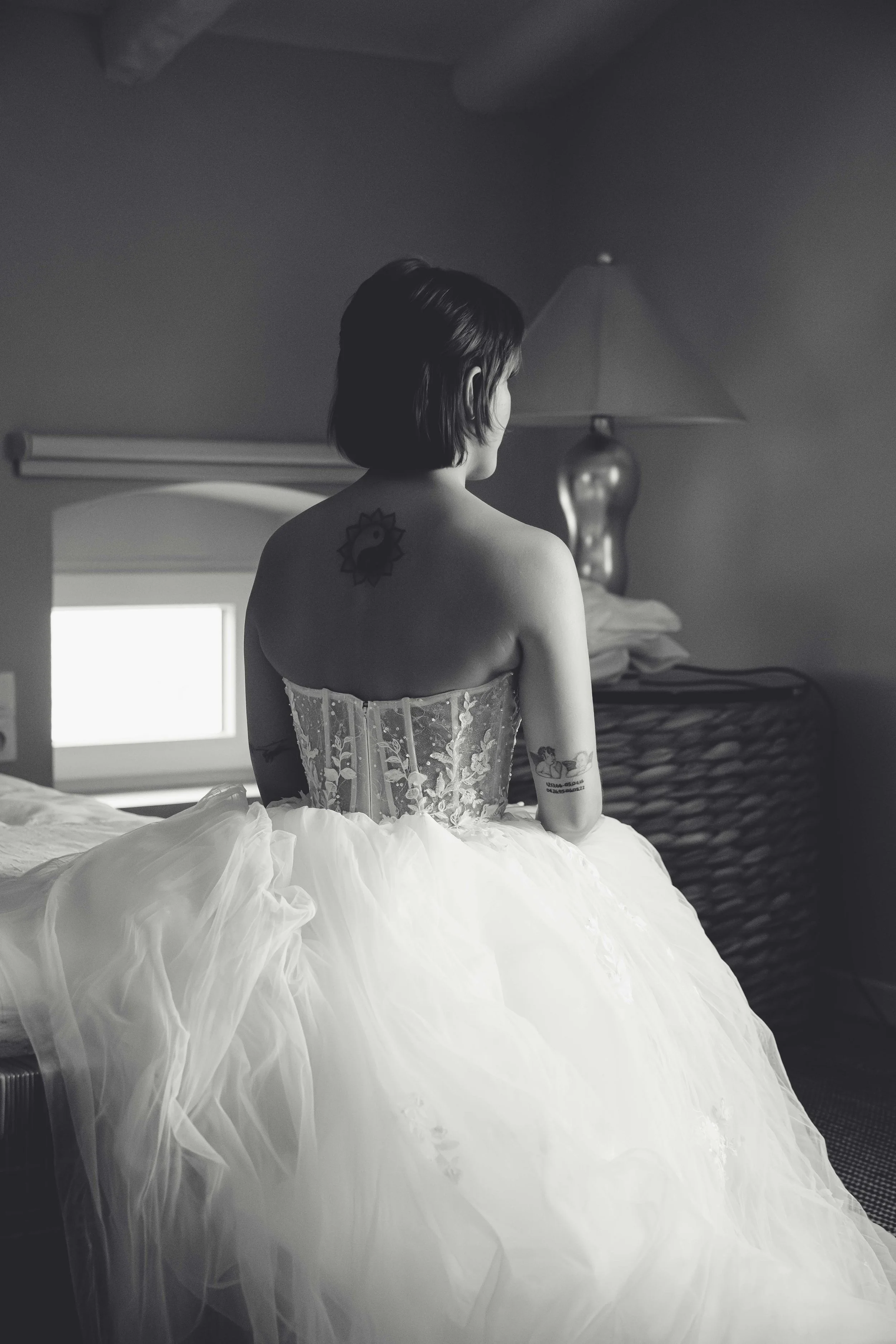 A woman with short hair and tattoos on her back, sitting on a bed, wearing a strapless wedding dress with a lace bodice and a voluminous tulle skirt, looking out of a small window in a dimly lit room.
