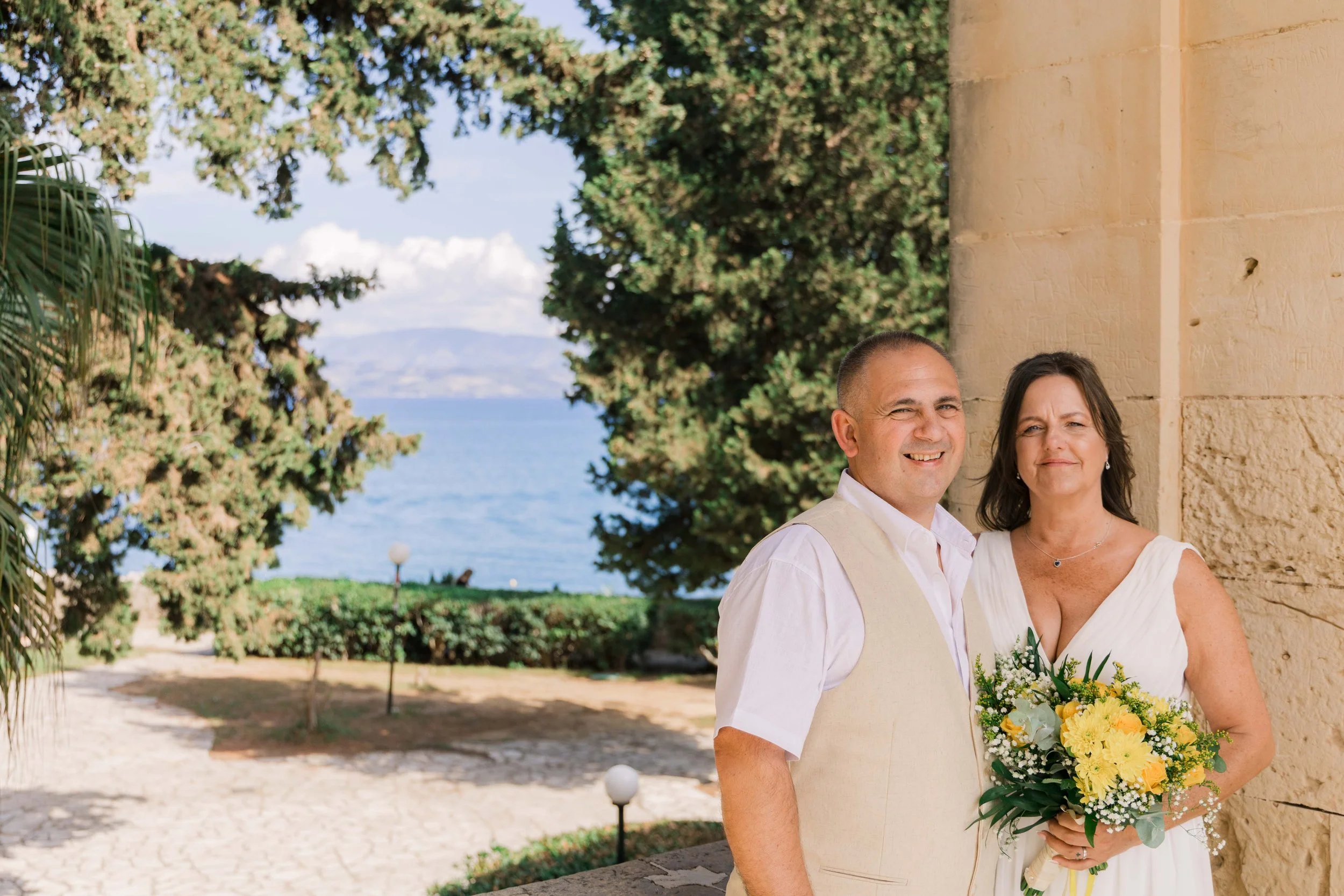A smiling man and woman stand outside near a stone building, with trees, a grass area, and a view of the ocean in the background. The woman holds a bouquet of yellow and white flowers.