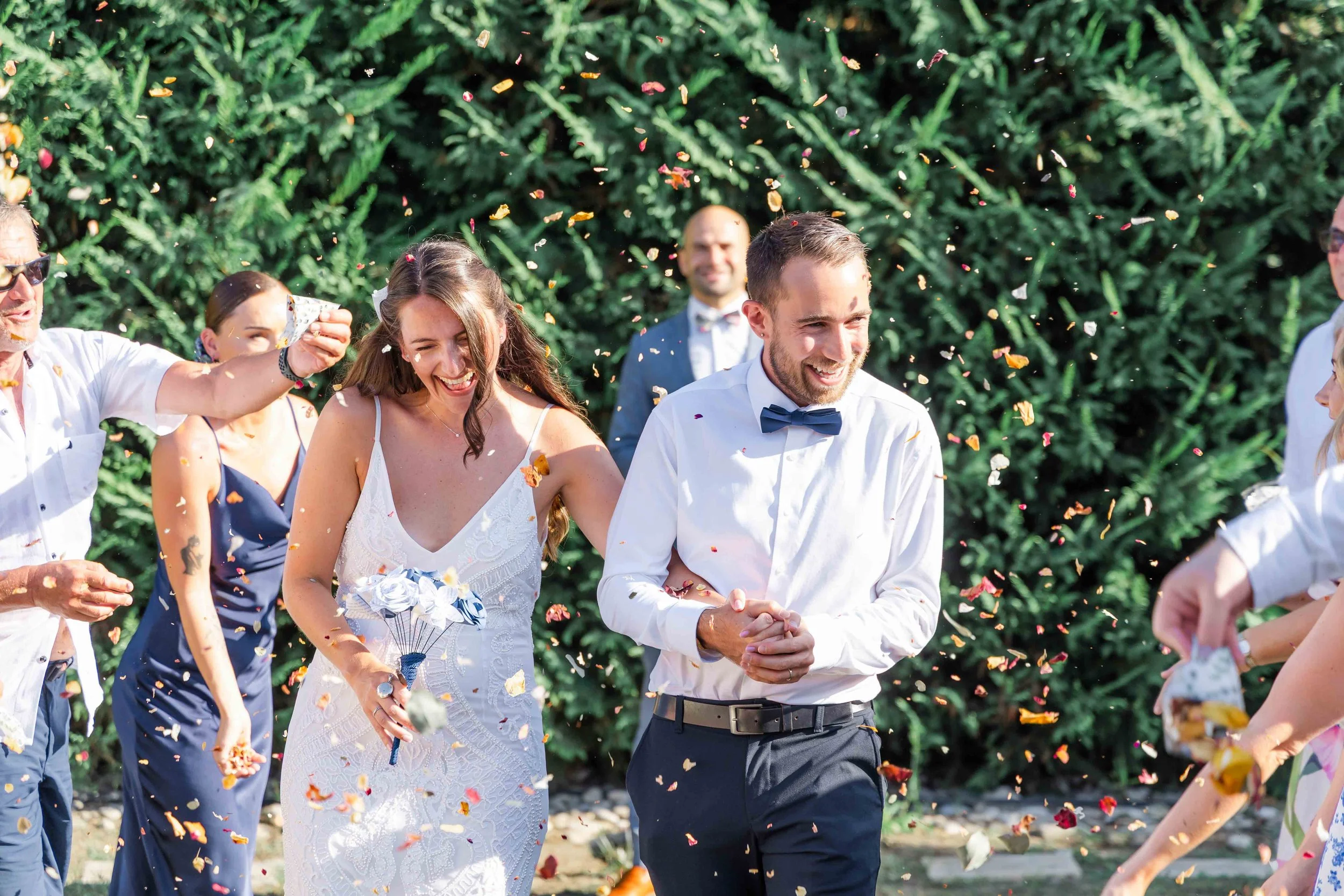 A wedding celebration outdoors with people throwing confetti, focusing on a bride and groom smiling and holding hands.
