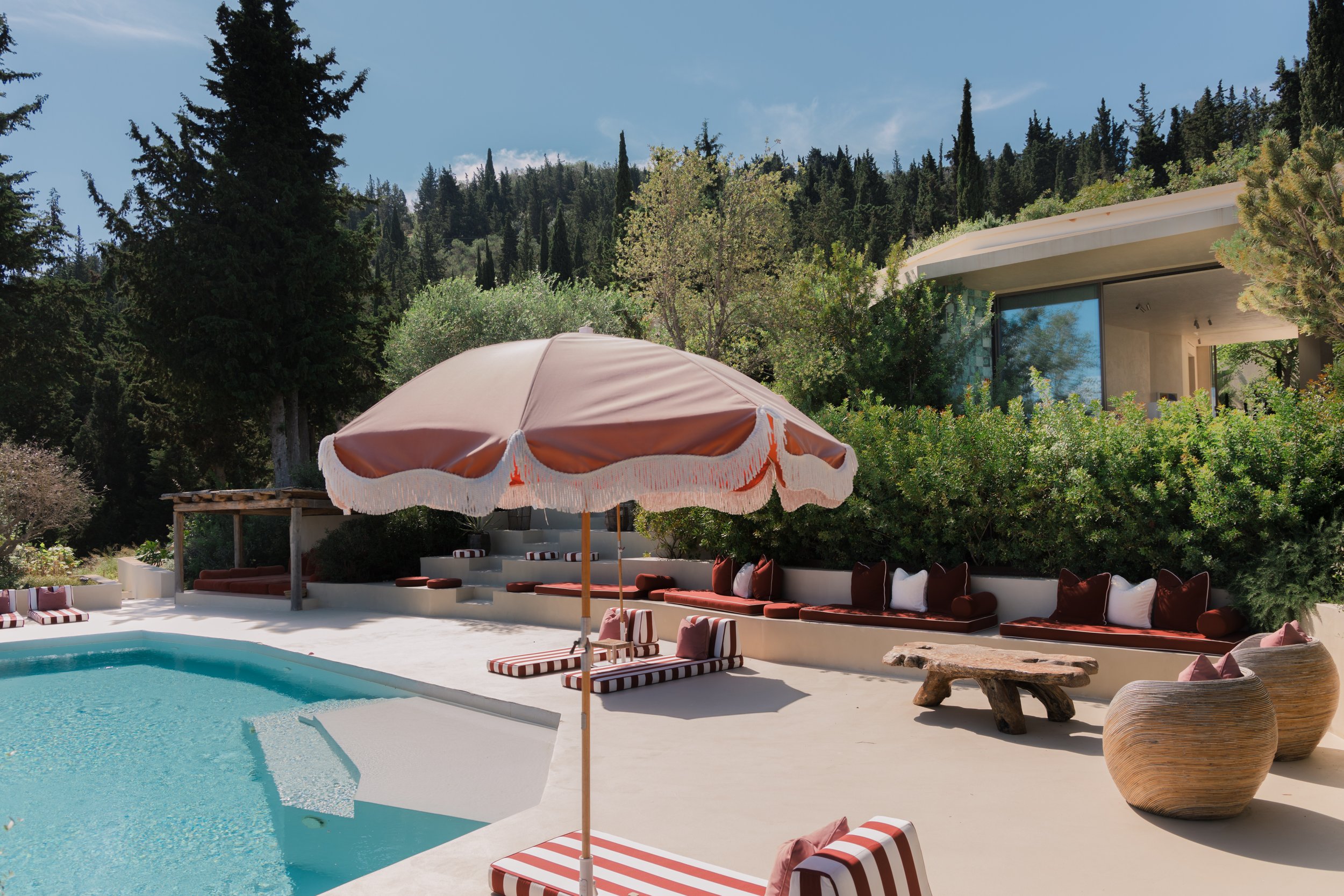 Luxury outdoor swimming pool area with poolside lounge chairs, striped umbrellas, and surrounding lush green trees and bushes, modern house with large glass windows in the background.