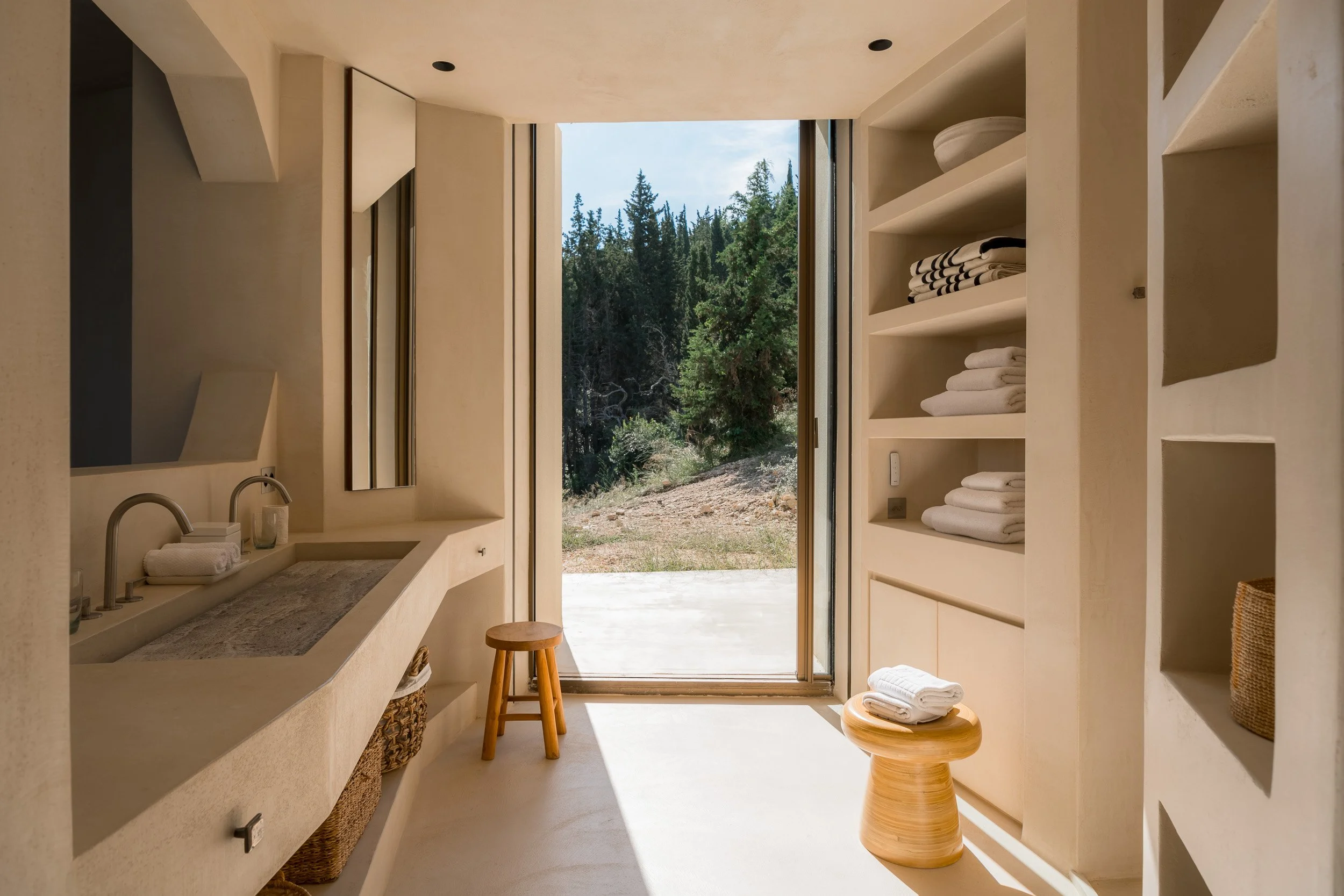 A minimalist bathroom with a built-in sink, open shelves with folded towels, a small wooden stool, and a large glass door opening to a natural outdoor scene with trees and blue sky.